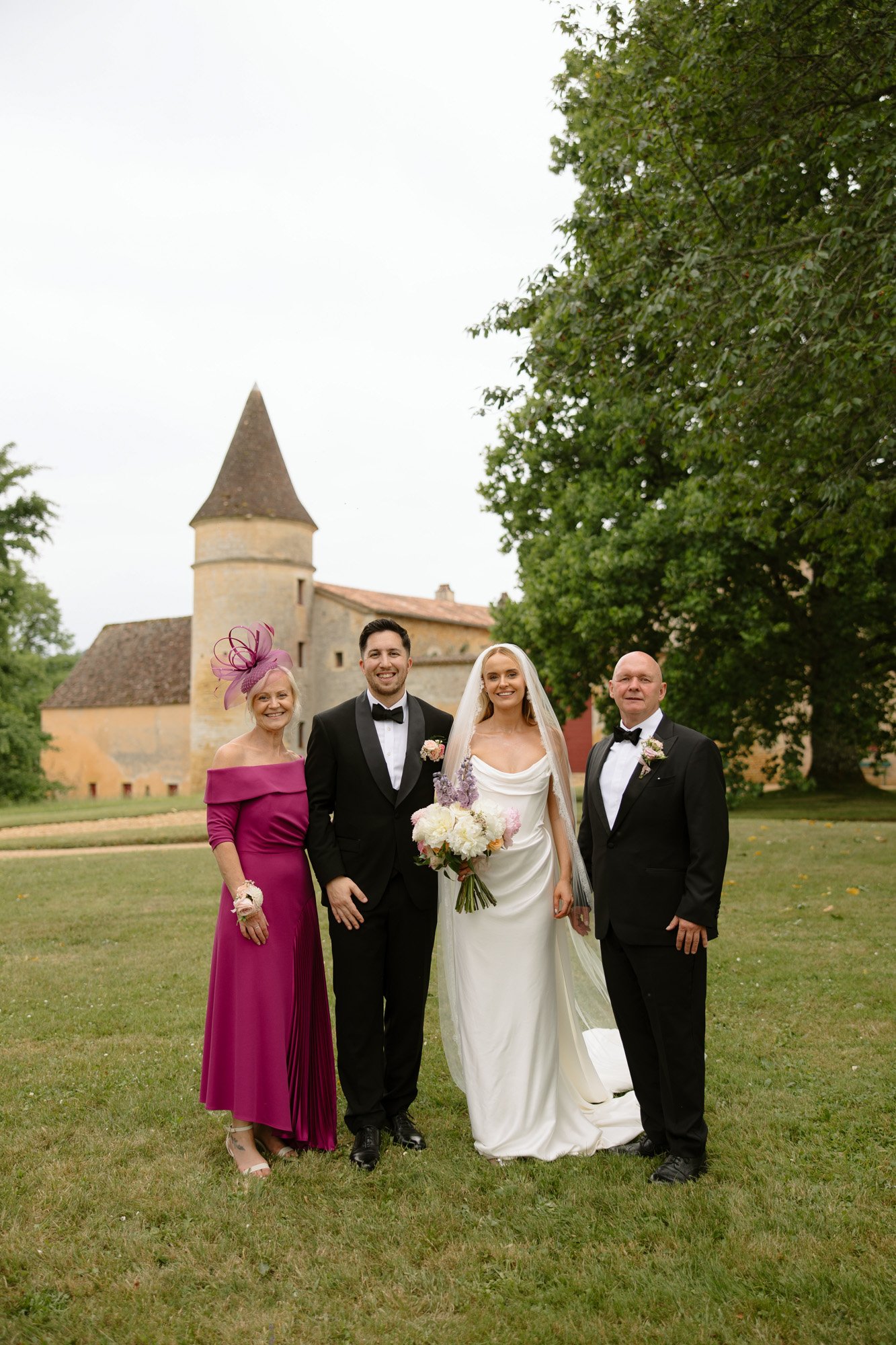 A bride and groom stand with two adults in formal attire on a lawn in front of a historic building with a tower. Chateau de la Bourlie wedding.