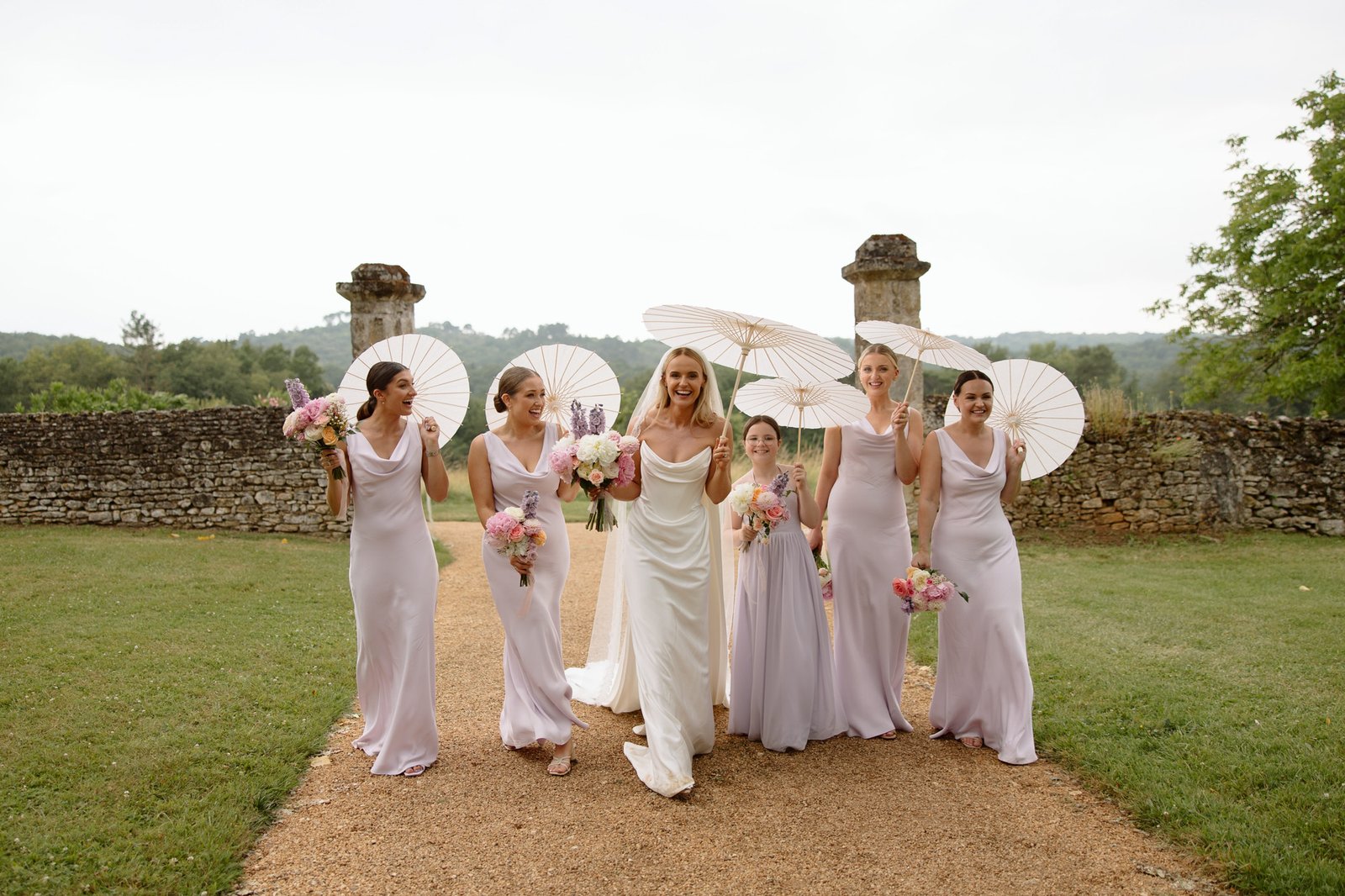 A bride in a white dress walks outdoors with five bridesmaids in matching light lavender dresses, holding flowers and white parasols, along a gravel path bordered by grass and stone walls. Chateau de la Bourlie wedding.
