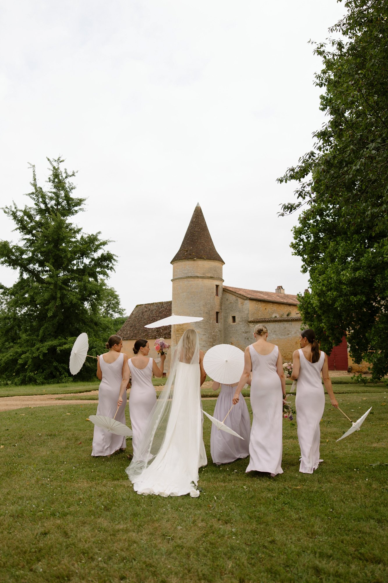 A bride in a long veil walks with five bridesmaids in matching light dresses, each holding a white parasol, near a stone building with a round tower. Chateau de la Bourlie wedding.