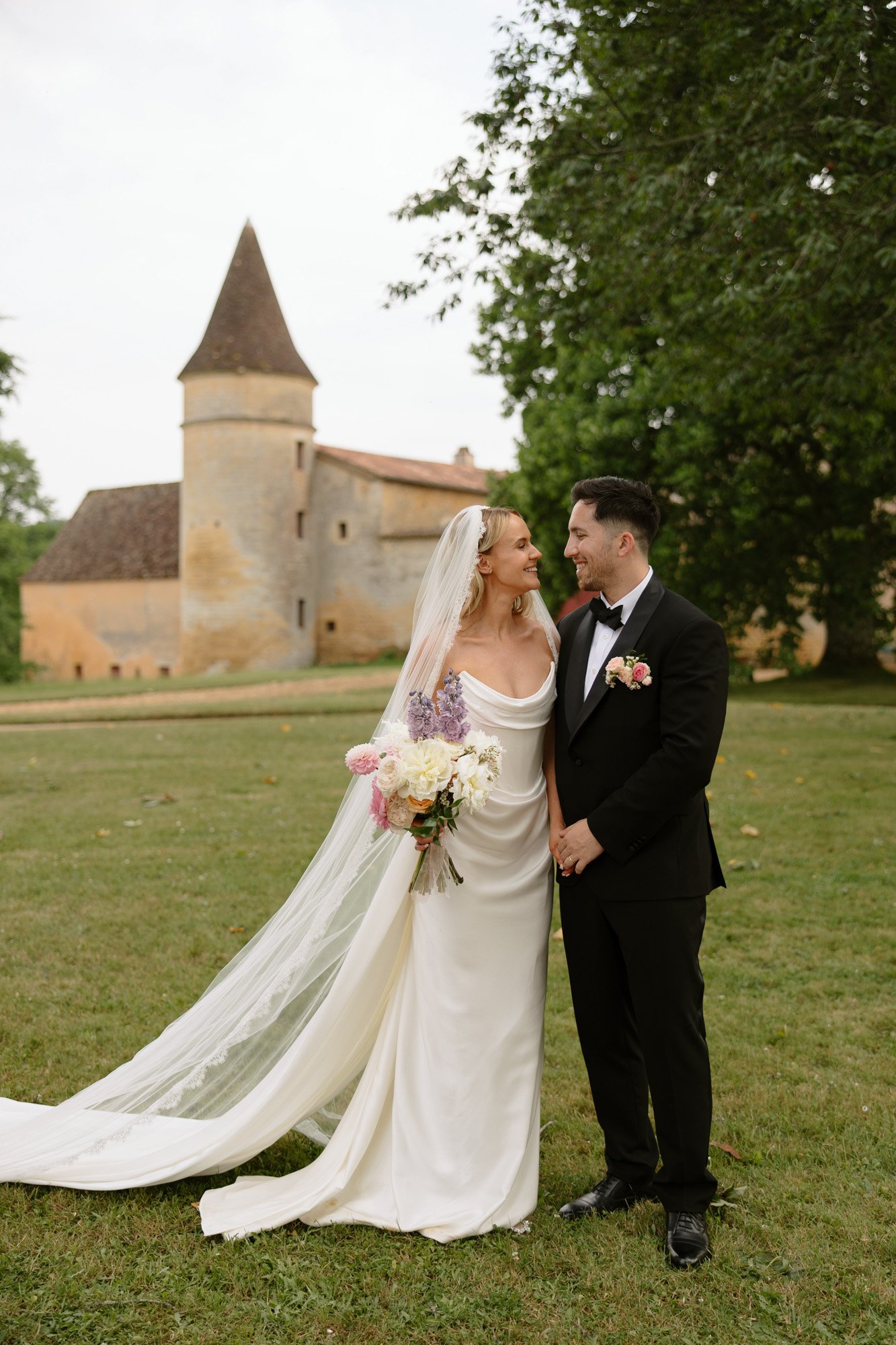 A bride in a white gown and veil holds a bouquet, standing beside a groom in a black tuxedo on grass, with a stone building and trees in the background. Chateau de la Bourlie wedding.