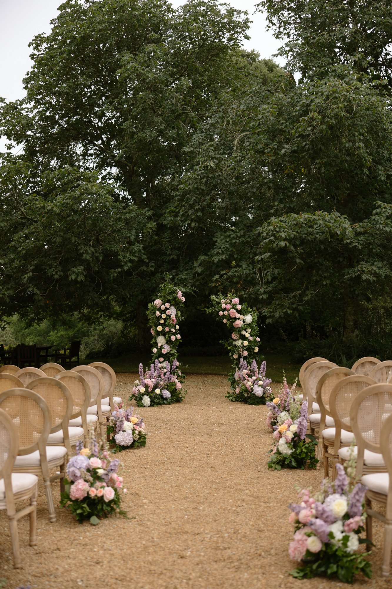 Outdoor wedding ceremony setup with rows of chairs facing a floral arch, surrounded by large trees and decorated with pastel flower arrangements along the aisle.