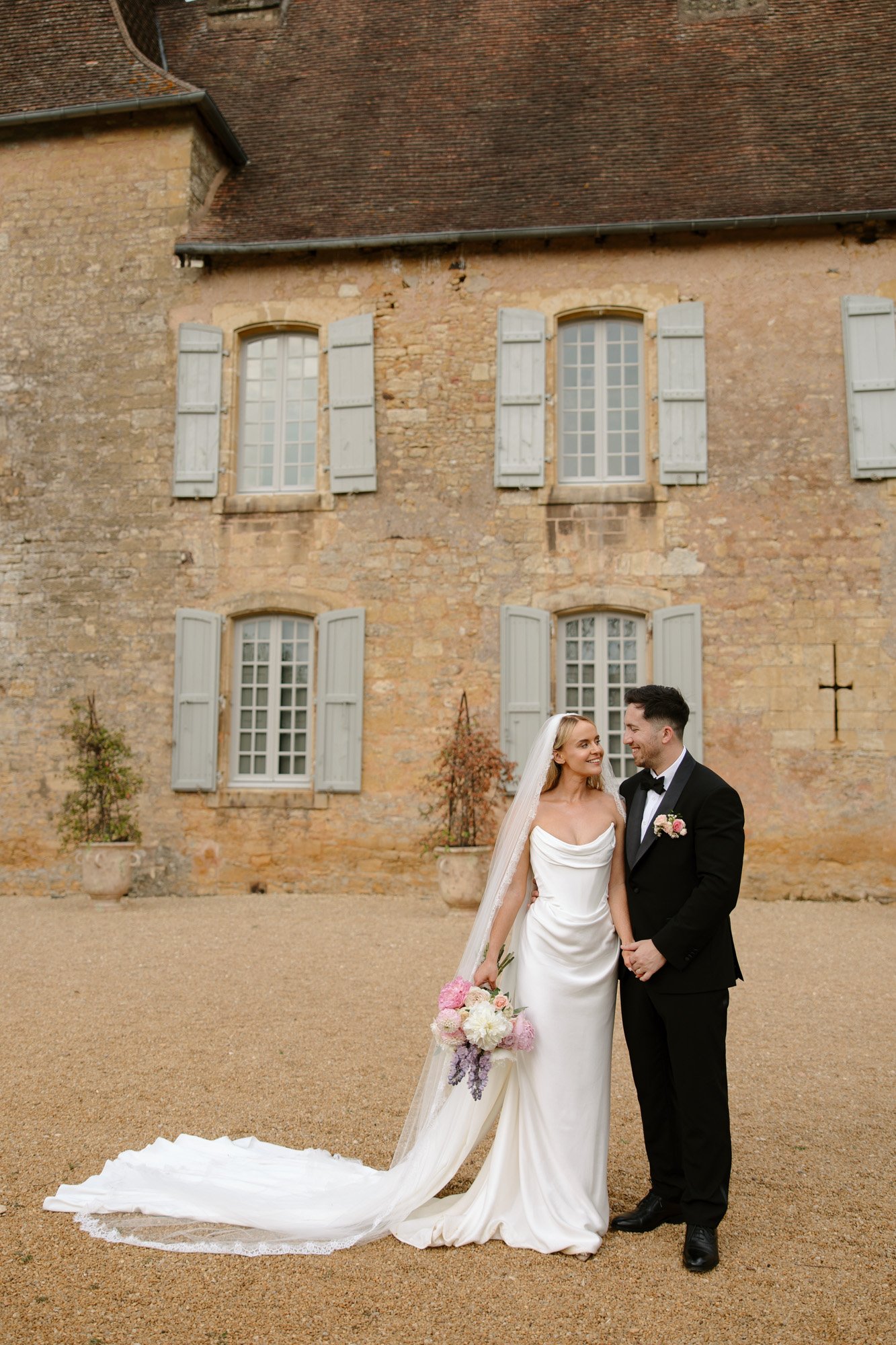A bride in a white dress and groom in a black tuxedo stand together outside a stone building with shuttered windows, smiling at each other.