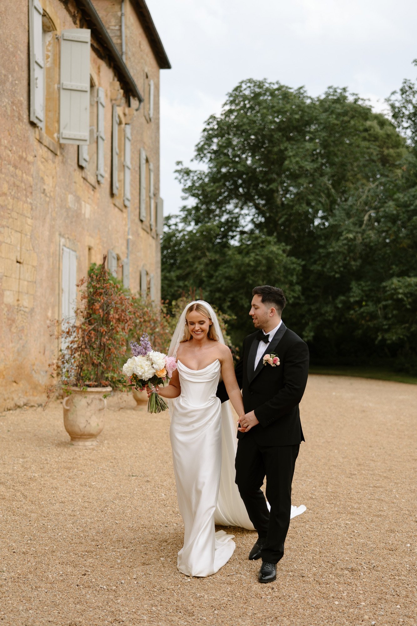 A bride in a white dress and veil holds a bouquet while walking outdoors with a groom in a black tuxedo beside a rustic building.