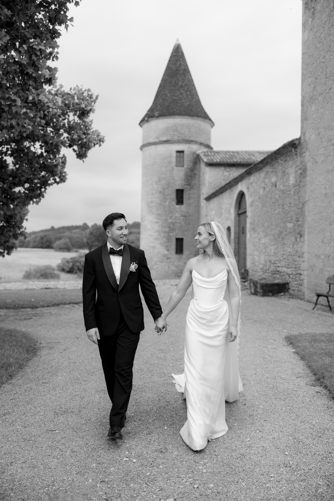 A bride and groom holding hands and walking outside a stone building with a tower, both dressed in formal wedding attire.
