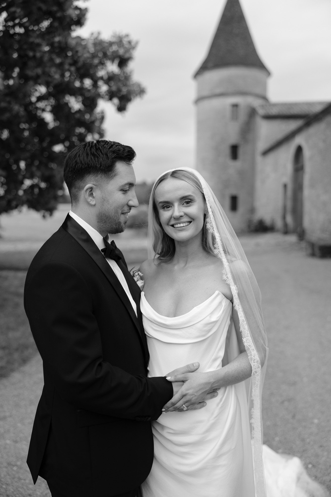 A bride and groom stand outdoors in front of a stone building with a tower. The bride wears a white gown and veil, and the groom is in a black tuxedo. Both are smiling.