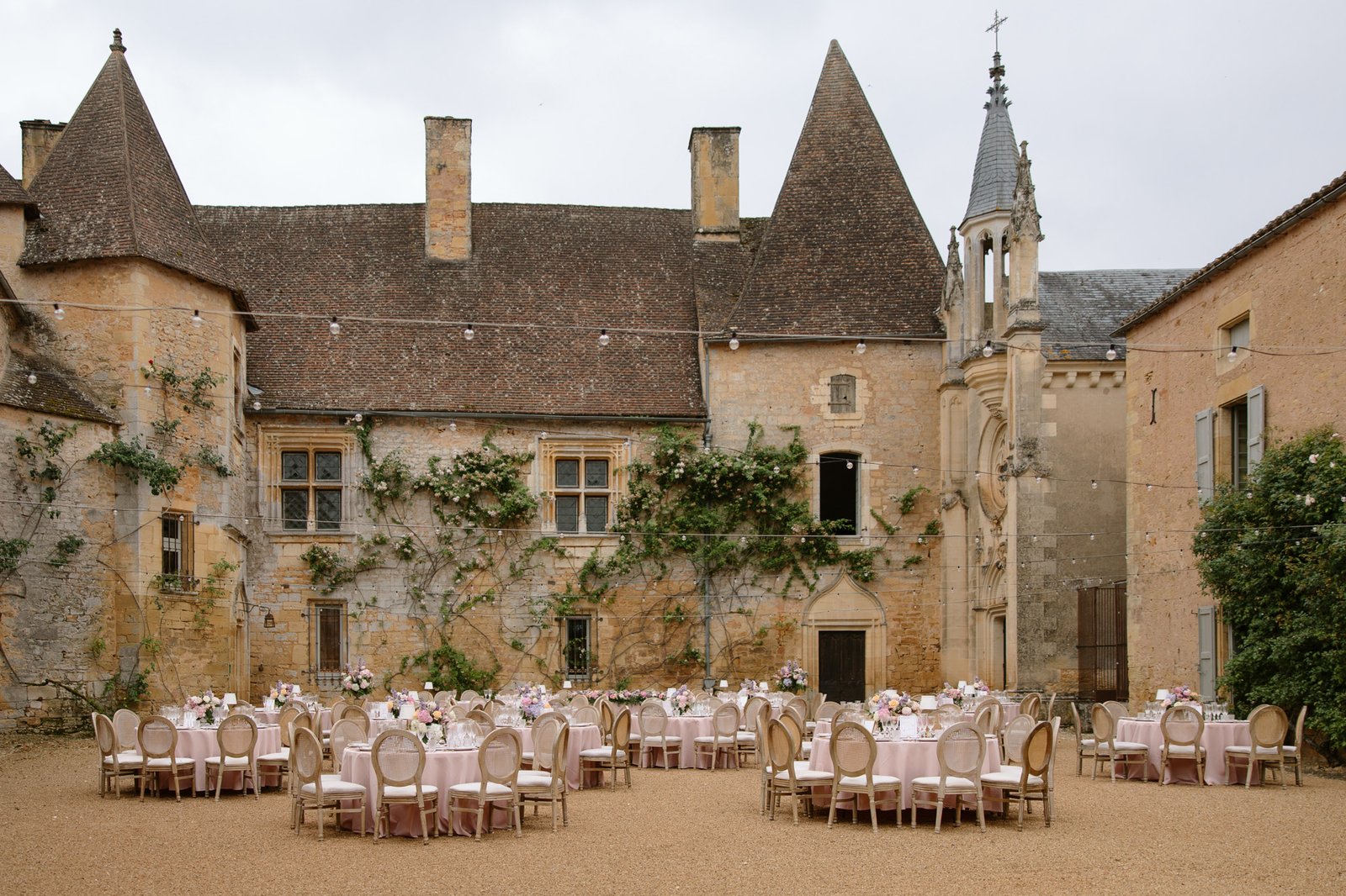Round tables with pink tablecloths and floral centerpieces are arranged in the courtyard of a historic stone building with ivy-covered walls and pointed rooftops. Chateau de la Bourlie wedding.