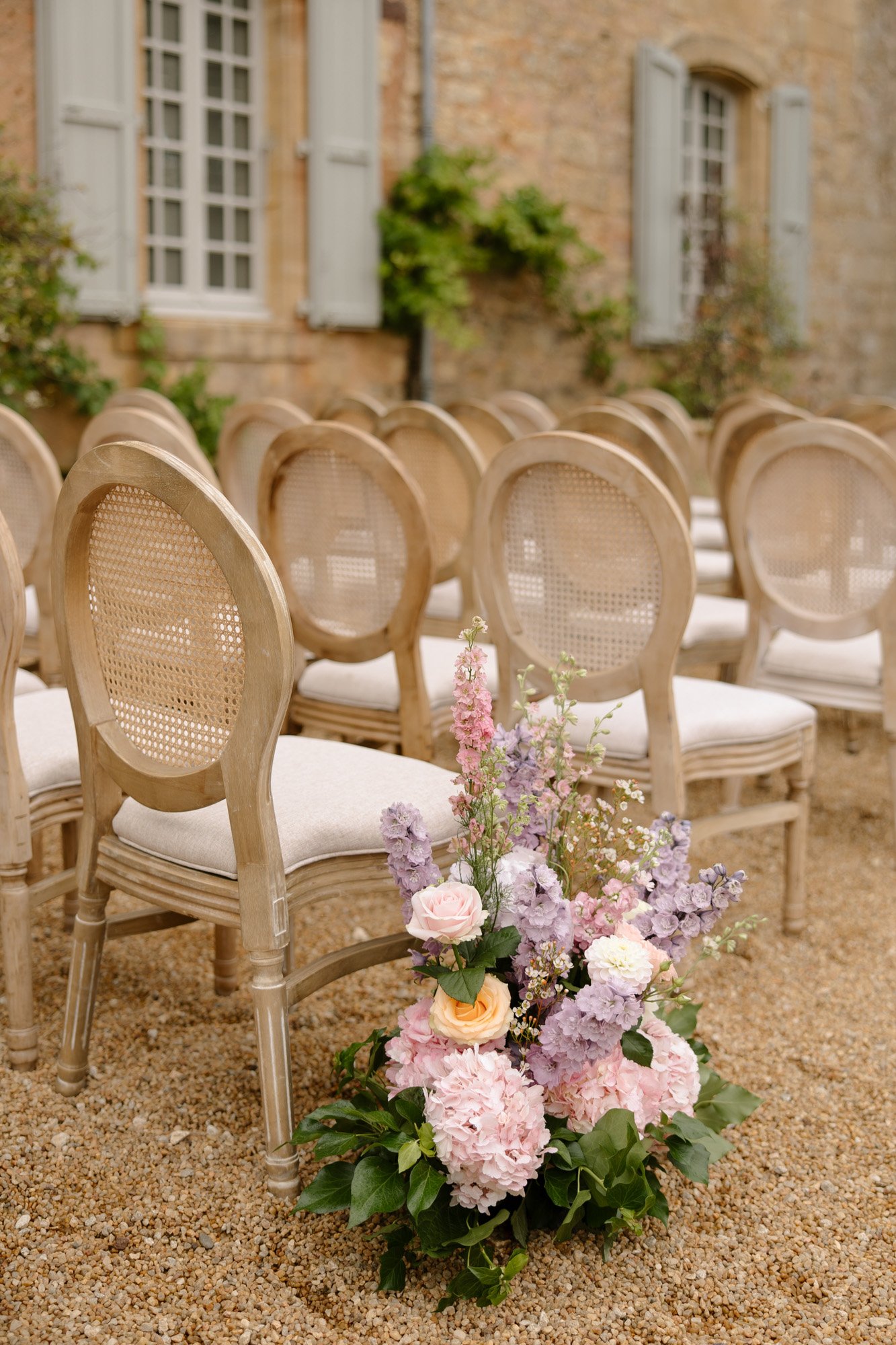 Rows of light wooden chairs with cream cushions are arranged outdoors on gravel, next to a large floral arrangement with pastel-colored flowers. A stone building with shuttered windows is in the background.