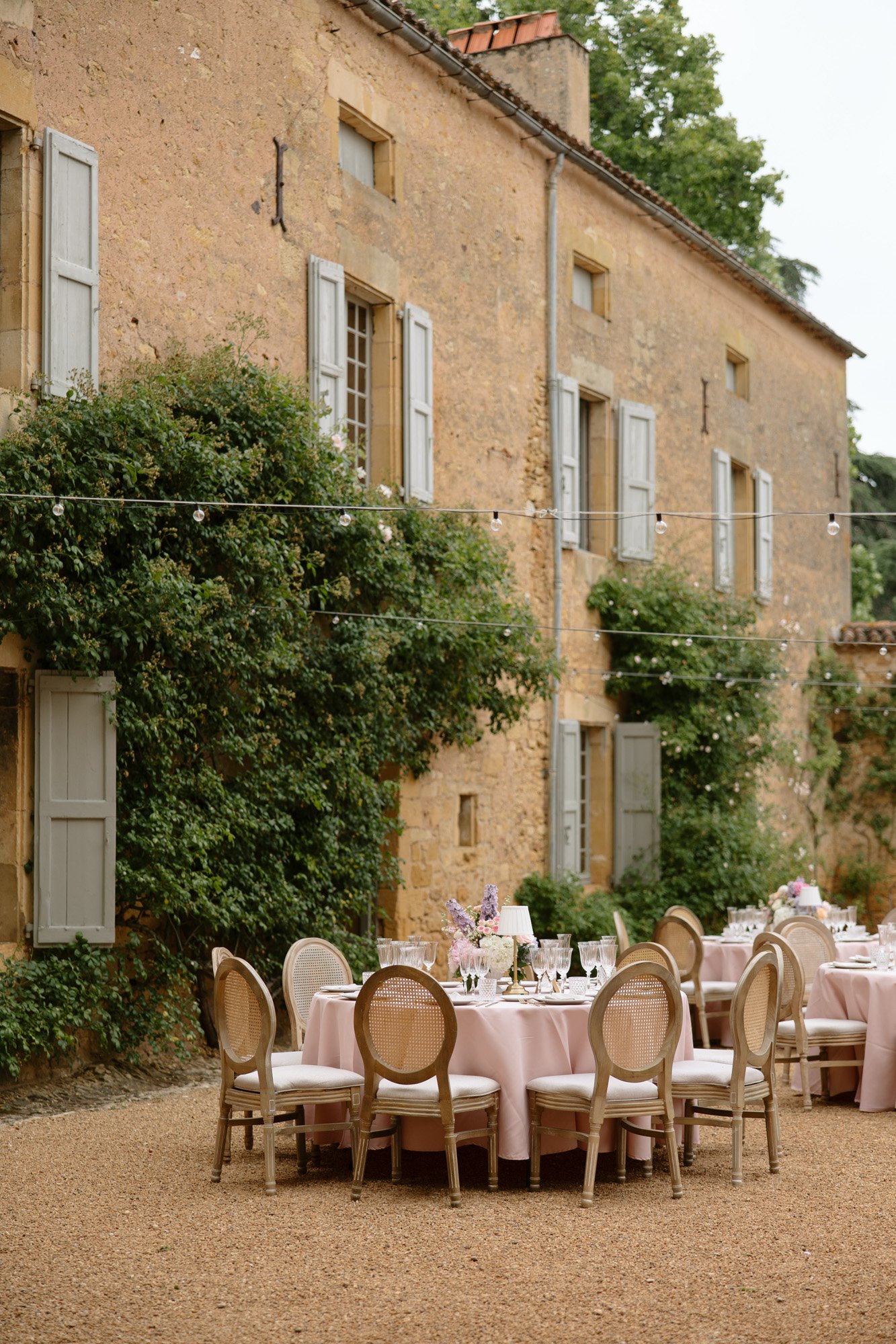 Outdoor dining tables with pink tablecloths and elegant chairs are set in front of an old stone building with ivy and shuttered windows, under string lights. Chateau de la Bourlie wedding.