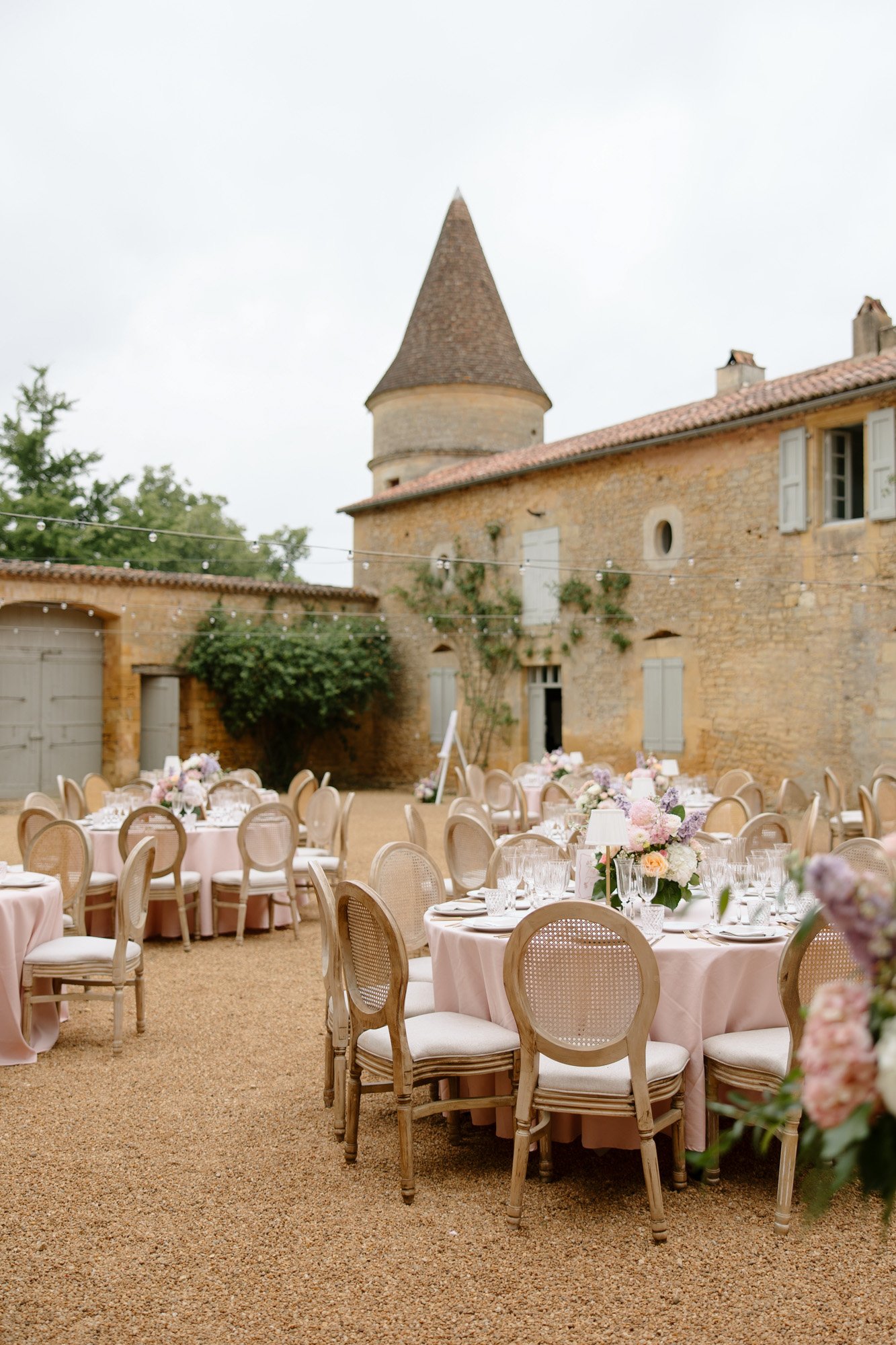 Outdoor courtyard set with round tables covered in pink tablecloths, surrounded by elegant chairs, floral centerpieces, and a stone building with a turret in the background.