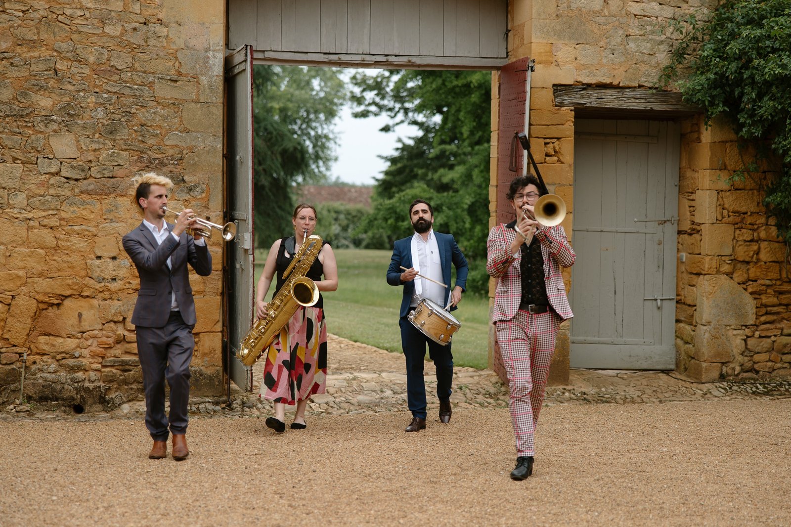 Four musicians play brass and percussion instruments while walking on a gravel path through an open stone gate in an outdoor setting.