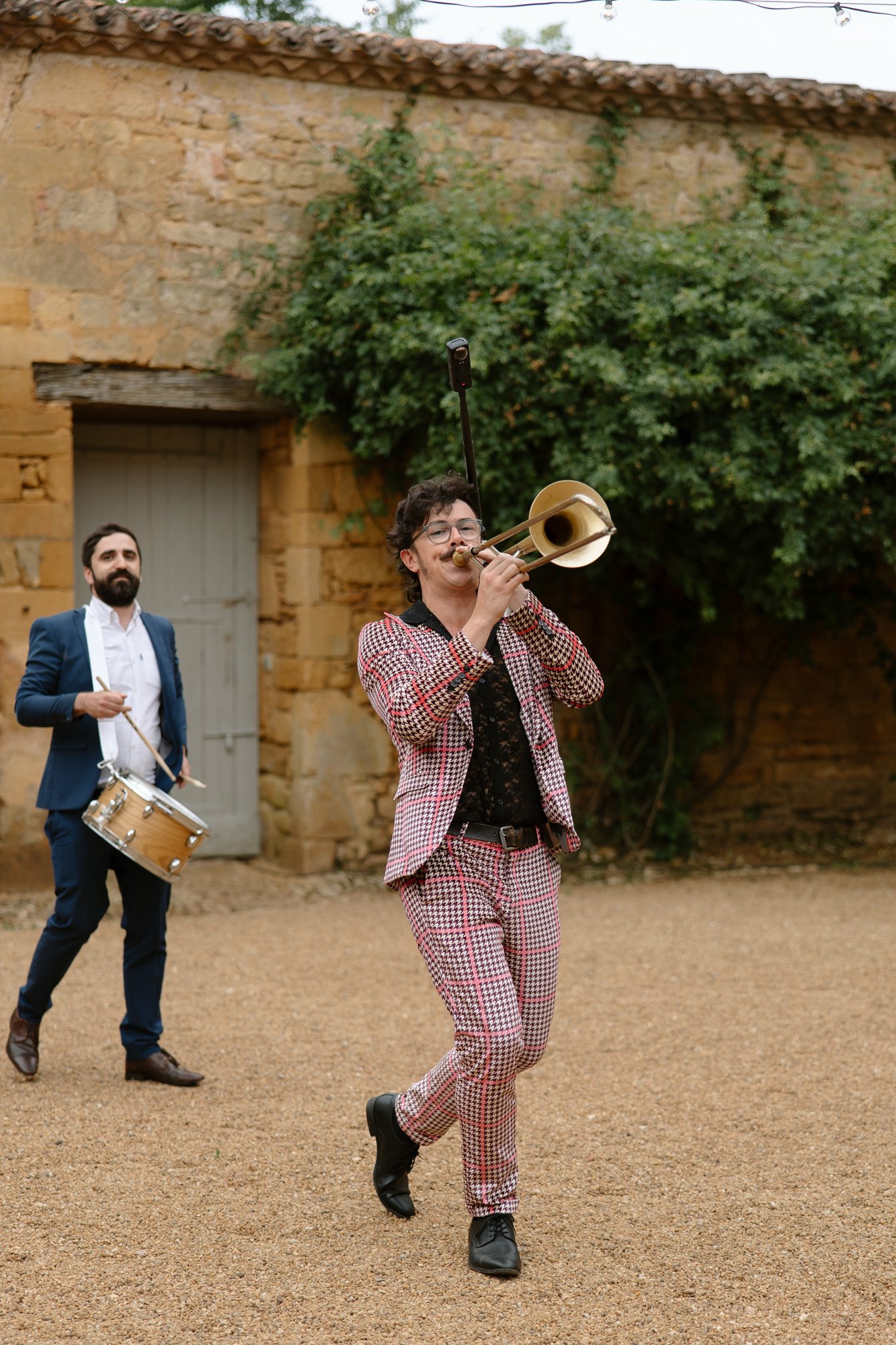 A man in a patterned suit plays the trombone while walking, followed by another man in a blue suit playing a snare drum, outdoors near a stone building.
