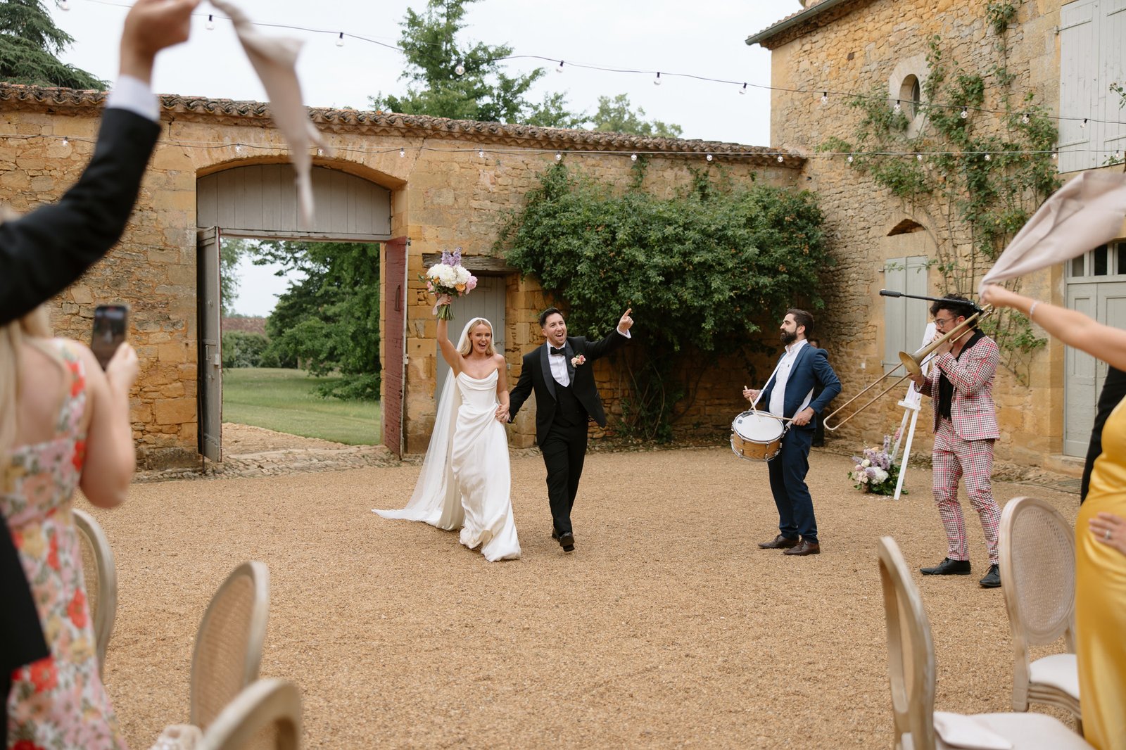 Bride and groom walk hand in hand outdoors as guests wave napkins and two musicians play drums and trombone in a courtyard with stone buildings. Chateau de la Bourlie wedding.