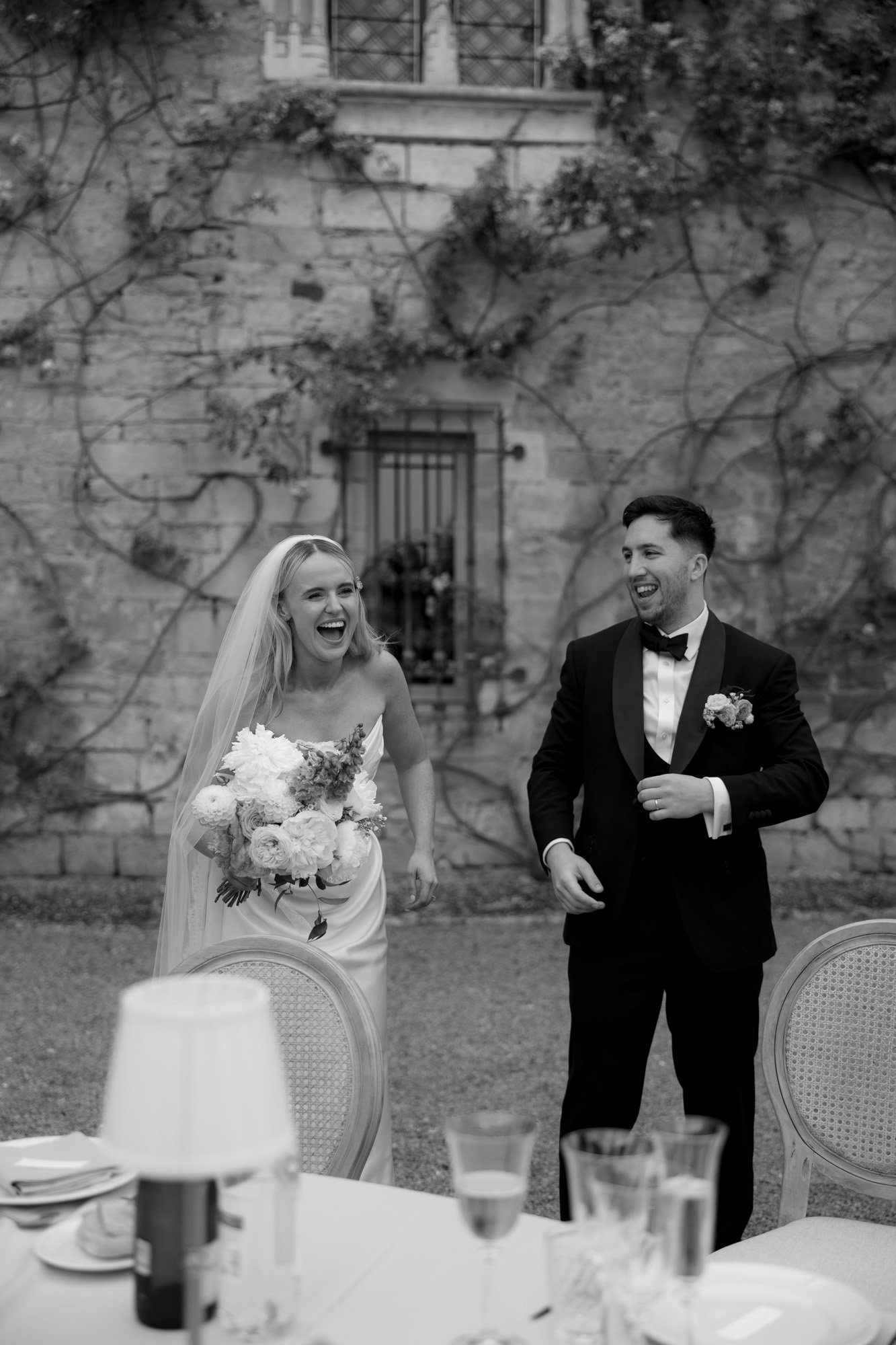 A bride holding a bouquet and a groom in a tuxedo smile and laugh outdoors near a stone wall with climbing vines.