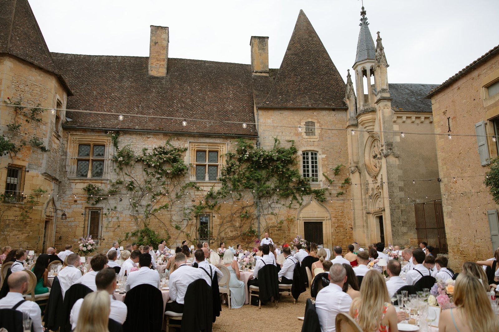 Guests seated at tables in a courtyard of a historic stone building during an outdoor formal event or wedding reception. Chateau de la Bourlie wedding.