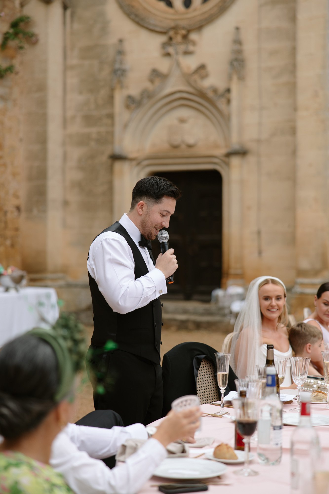 A man in a vest speaks into a microphone at an outdoor event, while guests, including a woman in a veil, sit at a table in front of a stone building.
