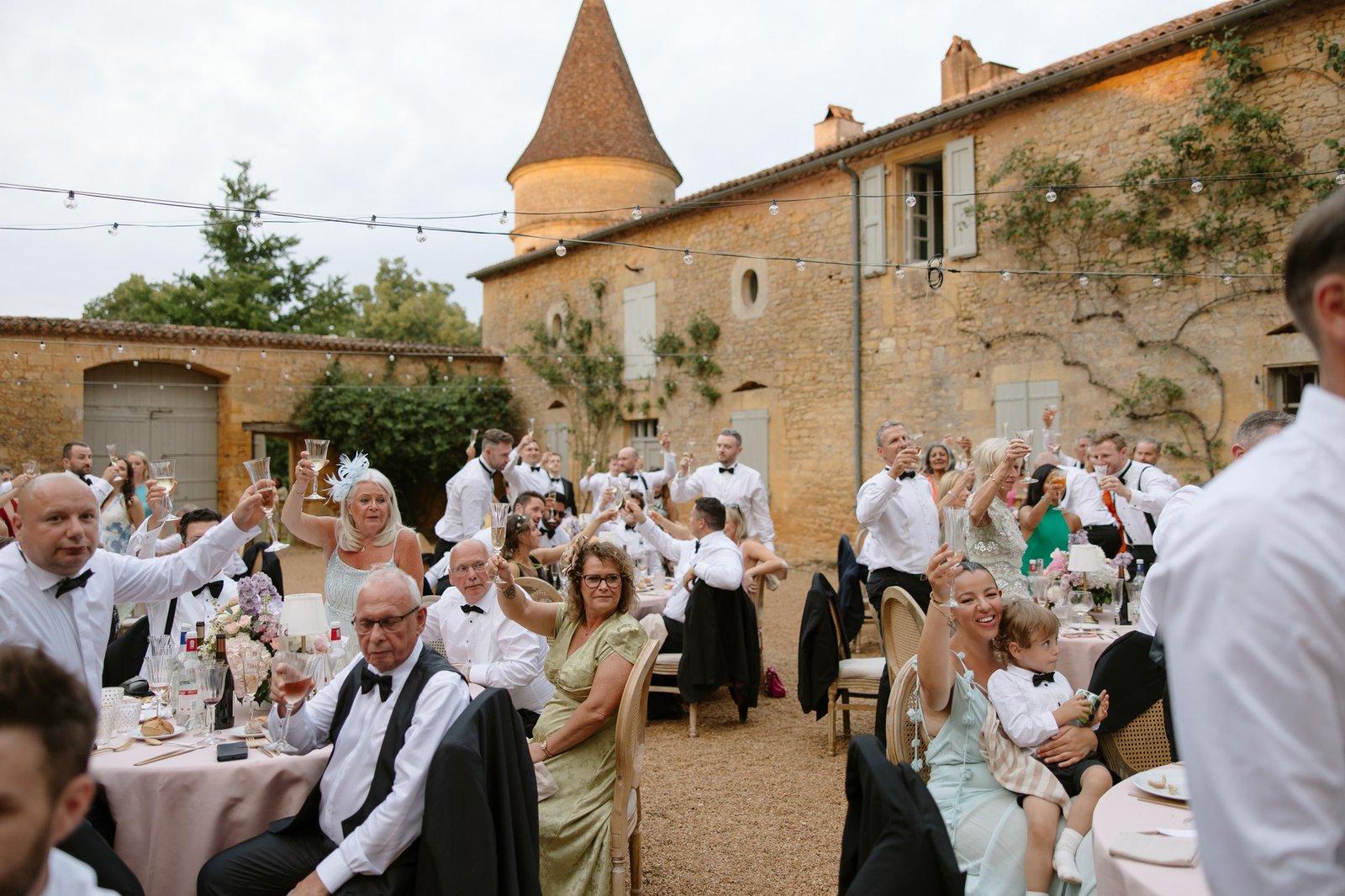 A group of people in formal attire raise glasses for a toast at an outdoor event in a courtyard with rustic stone buildings.