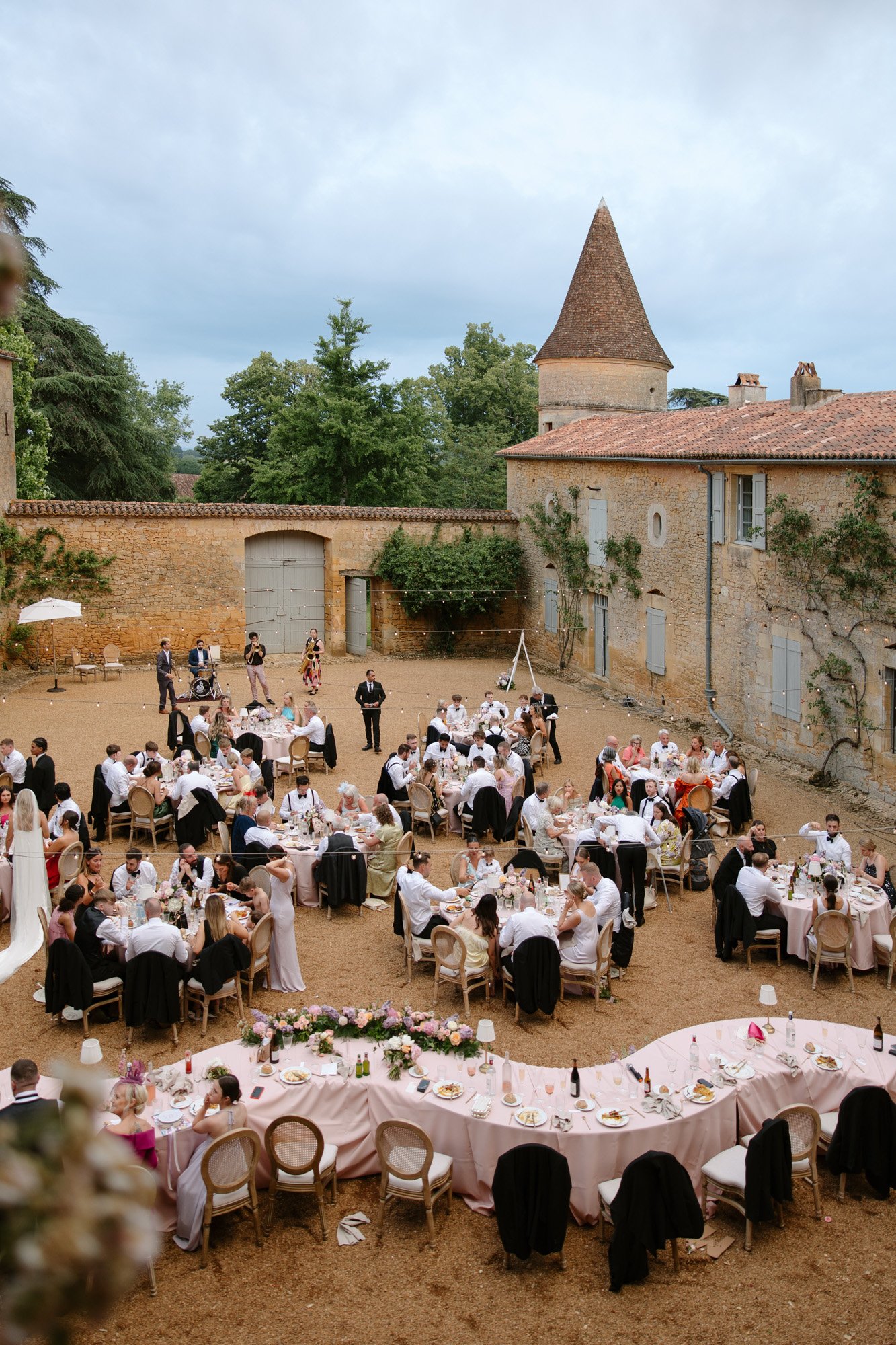 Outdoor wedding reception with guests seated at round and rectangular tables covered in pink tablecloths, set in a courtyard surrounded by rustic buildings. Chateau de la Bourlie wedding.