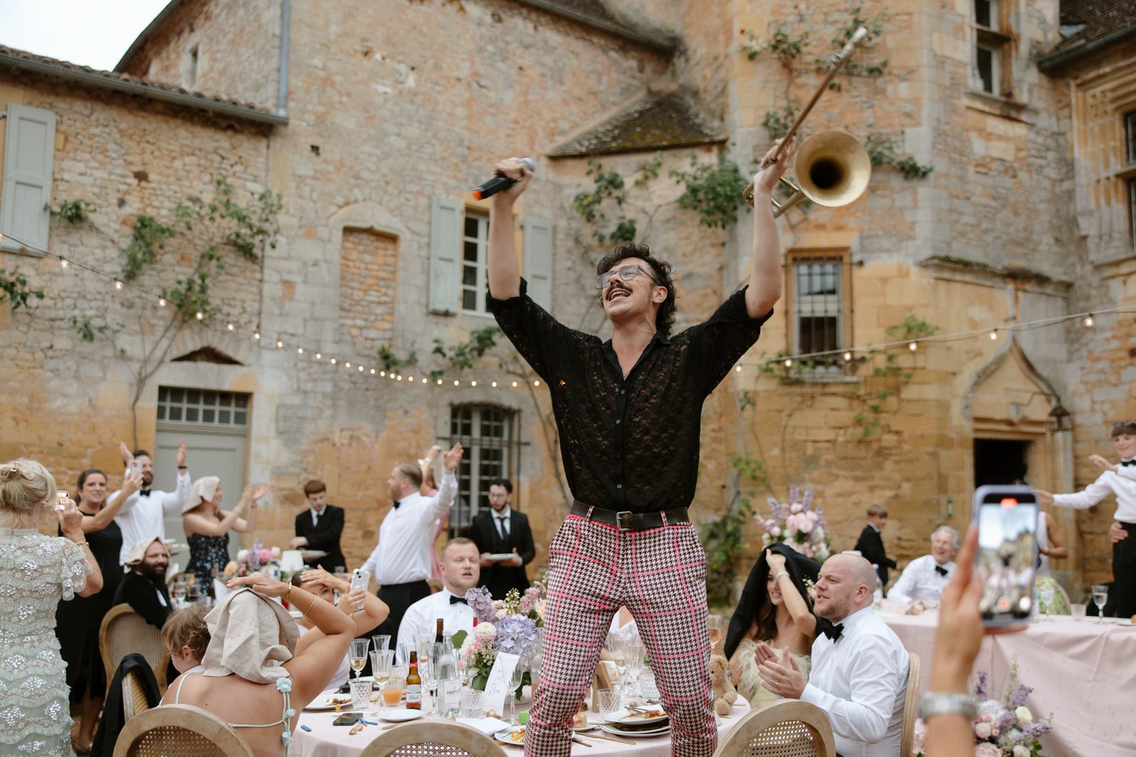A man stands on a table holding a microphone and trumpet, celebrating at an outdoor event with guests seated around him in front of a historic stone building.