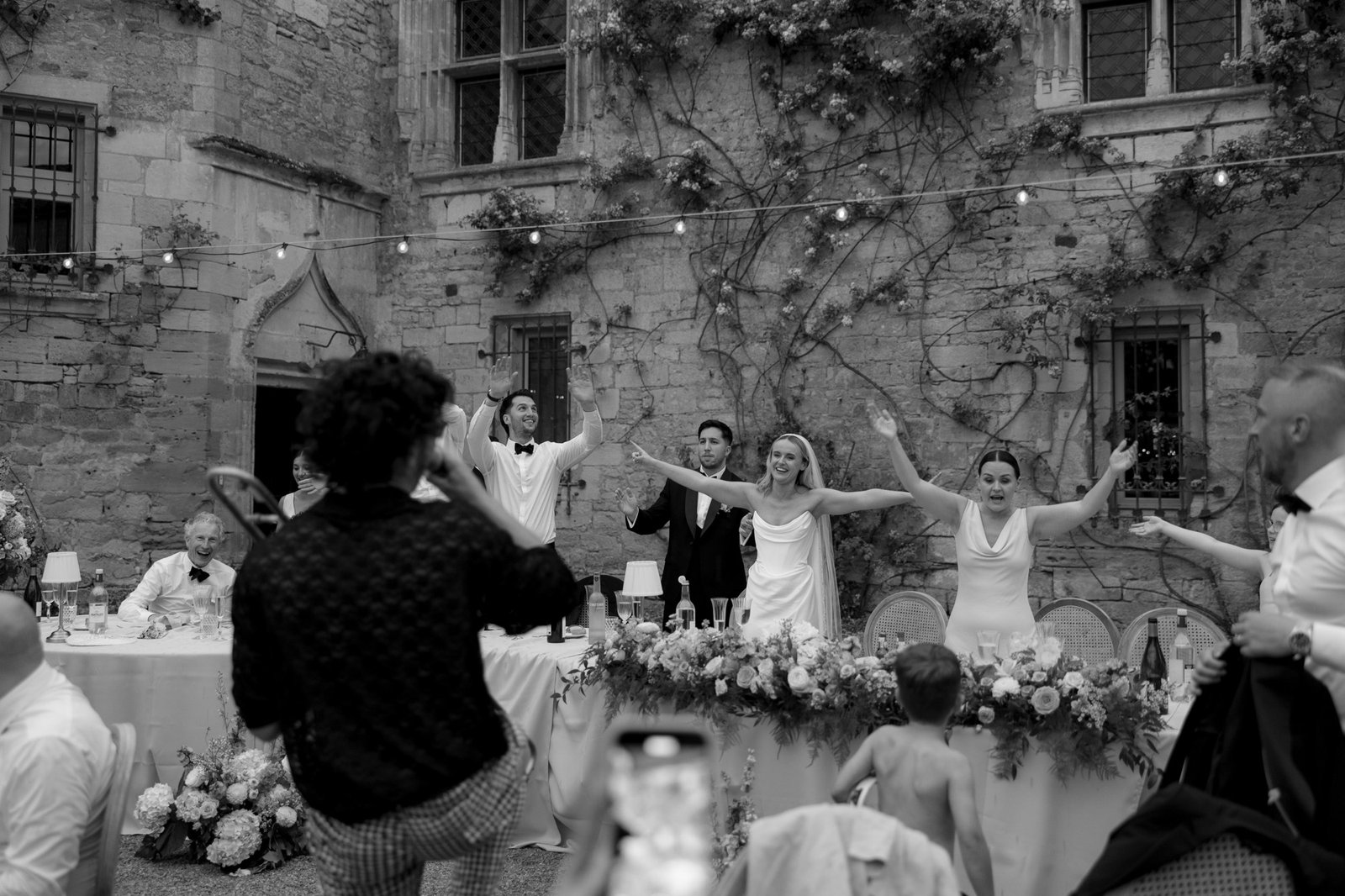 A wedding party stands and raises their arms in celebration behind a decorated table outdoors, while a photographer captures the moment. Guests and string lights are visible.