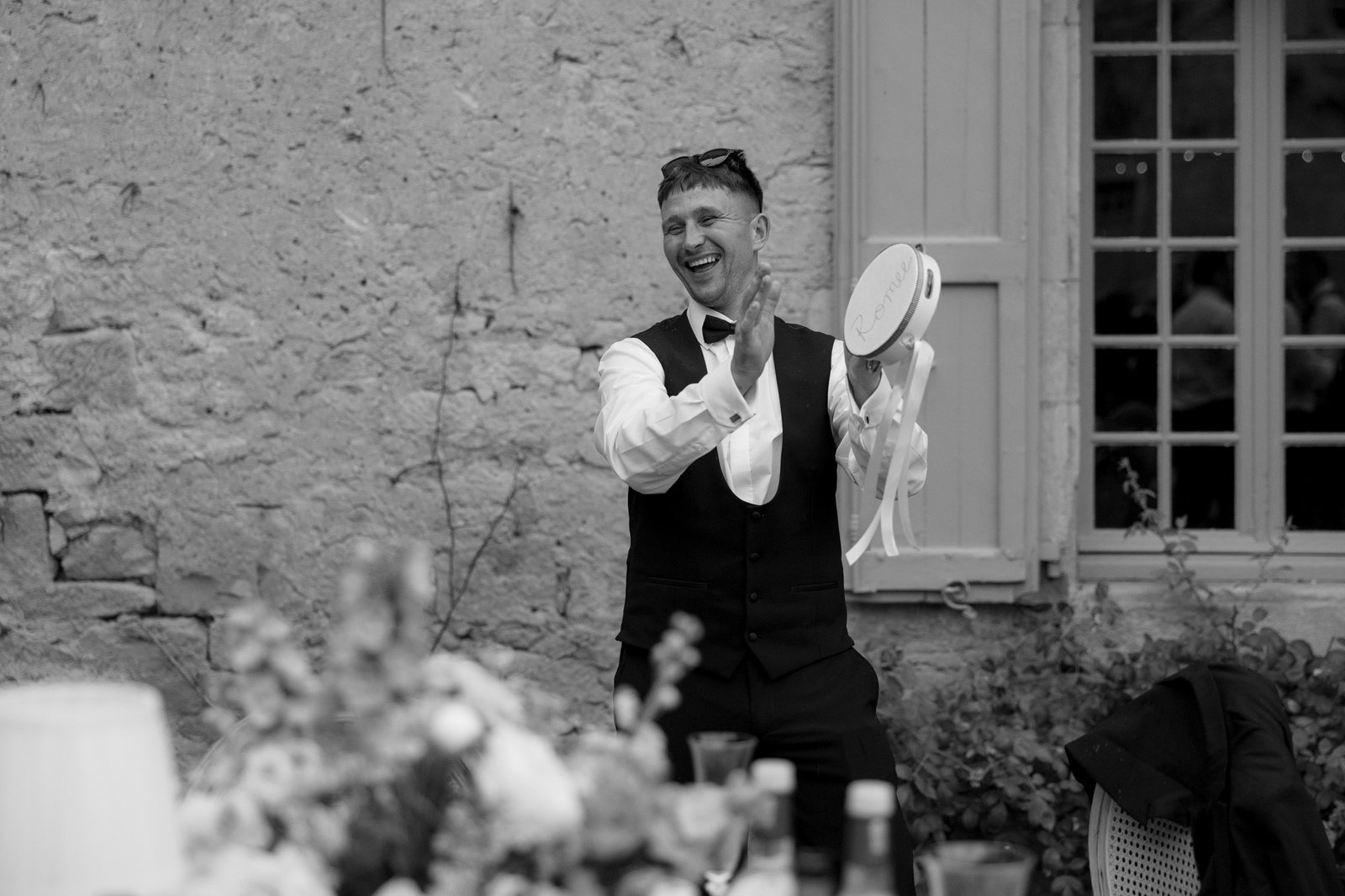 A man in formal attire stands indoors, holding a tambourine and clapping, with a table and blurred flowers in the foreground.