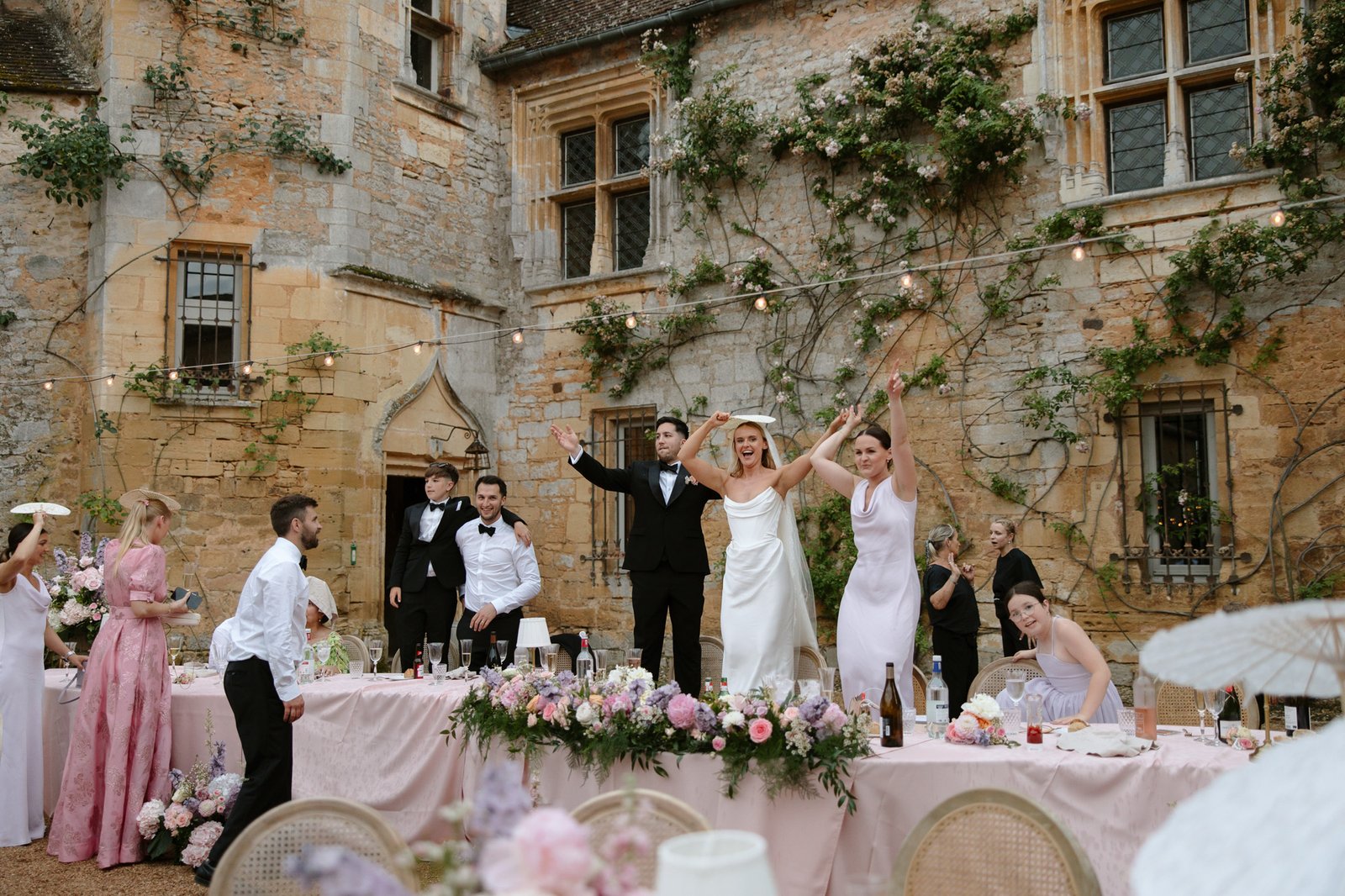 A bride, groom, and guests stand and celebrate on tables decorated with pink flowers at an outdoor wedding reception beside an old stone building. Chateau de la Bourlie wedding.
