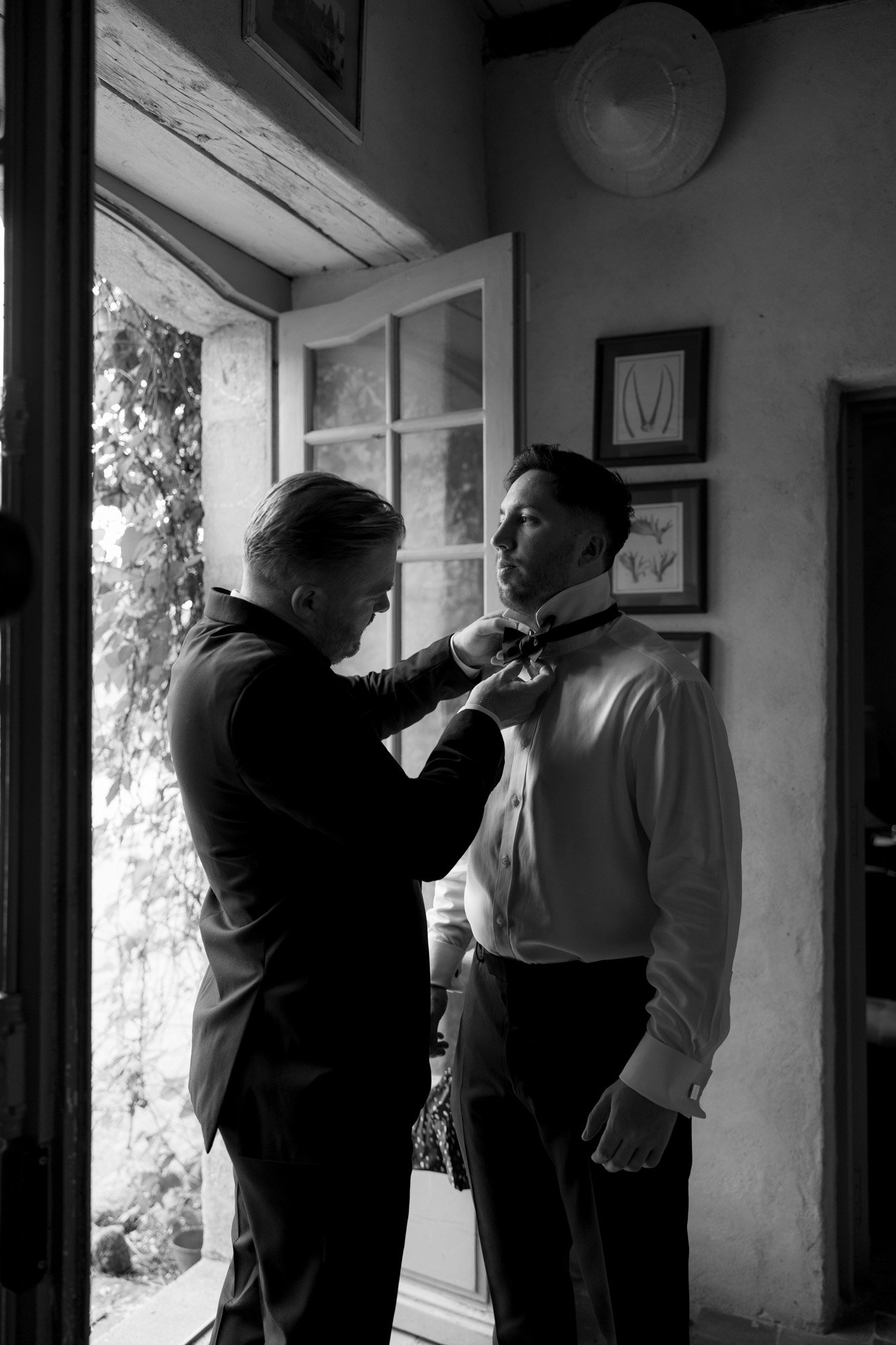 A man in a suit helps another man adjust his bow tie in a doorway, both wearing formal attire in a softly lit room. Chateau de la Bourlie wedding.