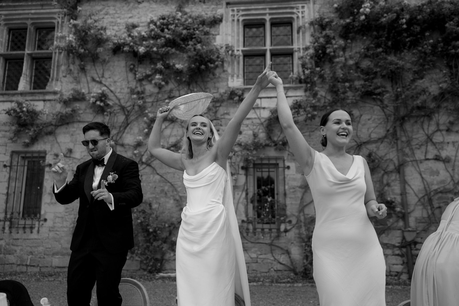 Three people dressed formally celebrate outdoors; two women in white dresses raise their hands together while a man in a suit stands beside them. A stone building with ivy is in the background.