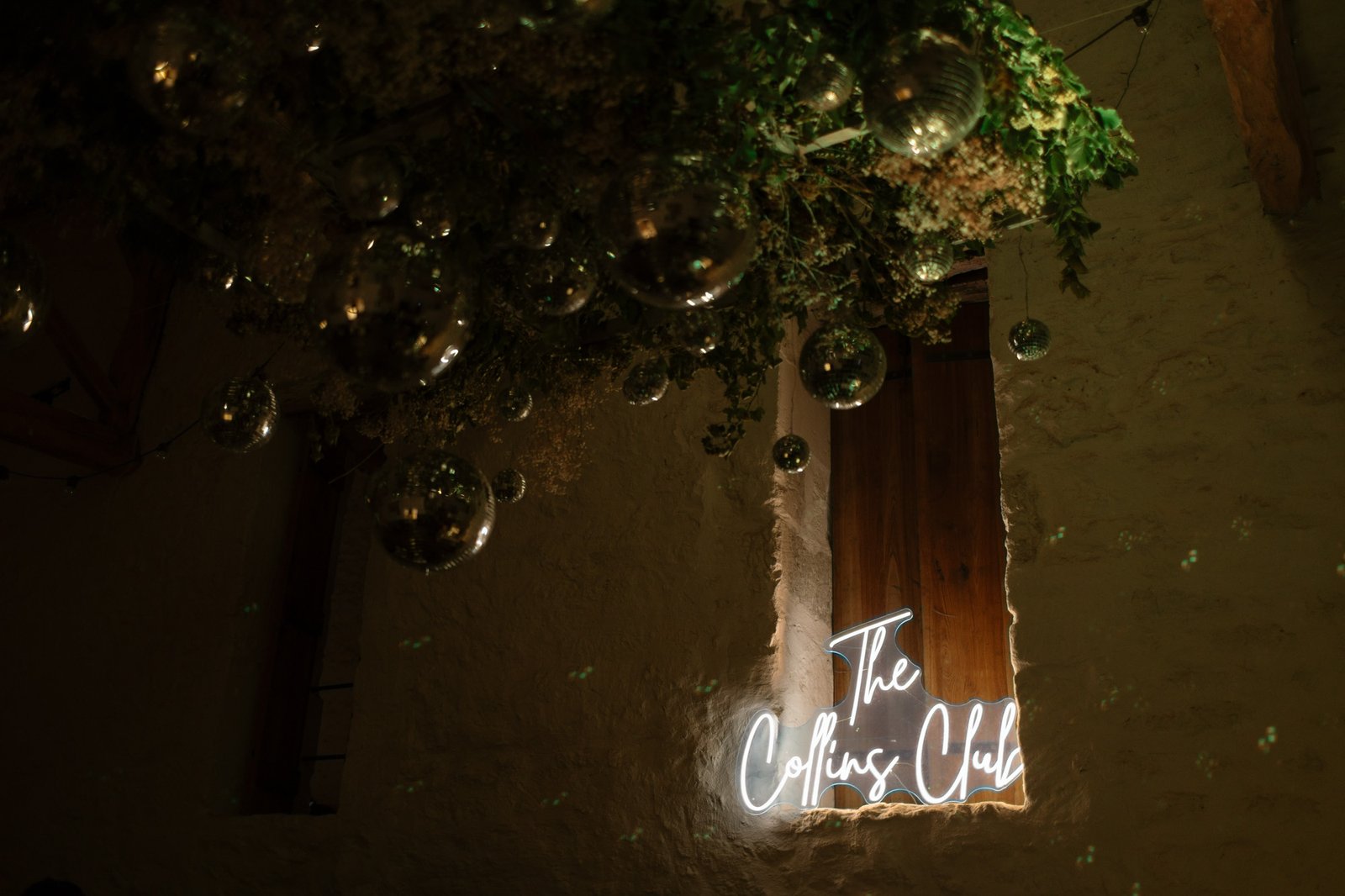 A neon sign reading The Collins Club is mounted on a stone wall under a wooden window, with hanging greenery and reflective orbs above.
