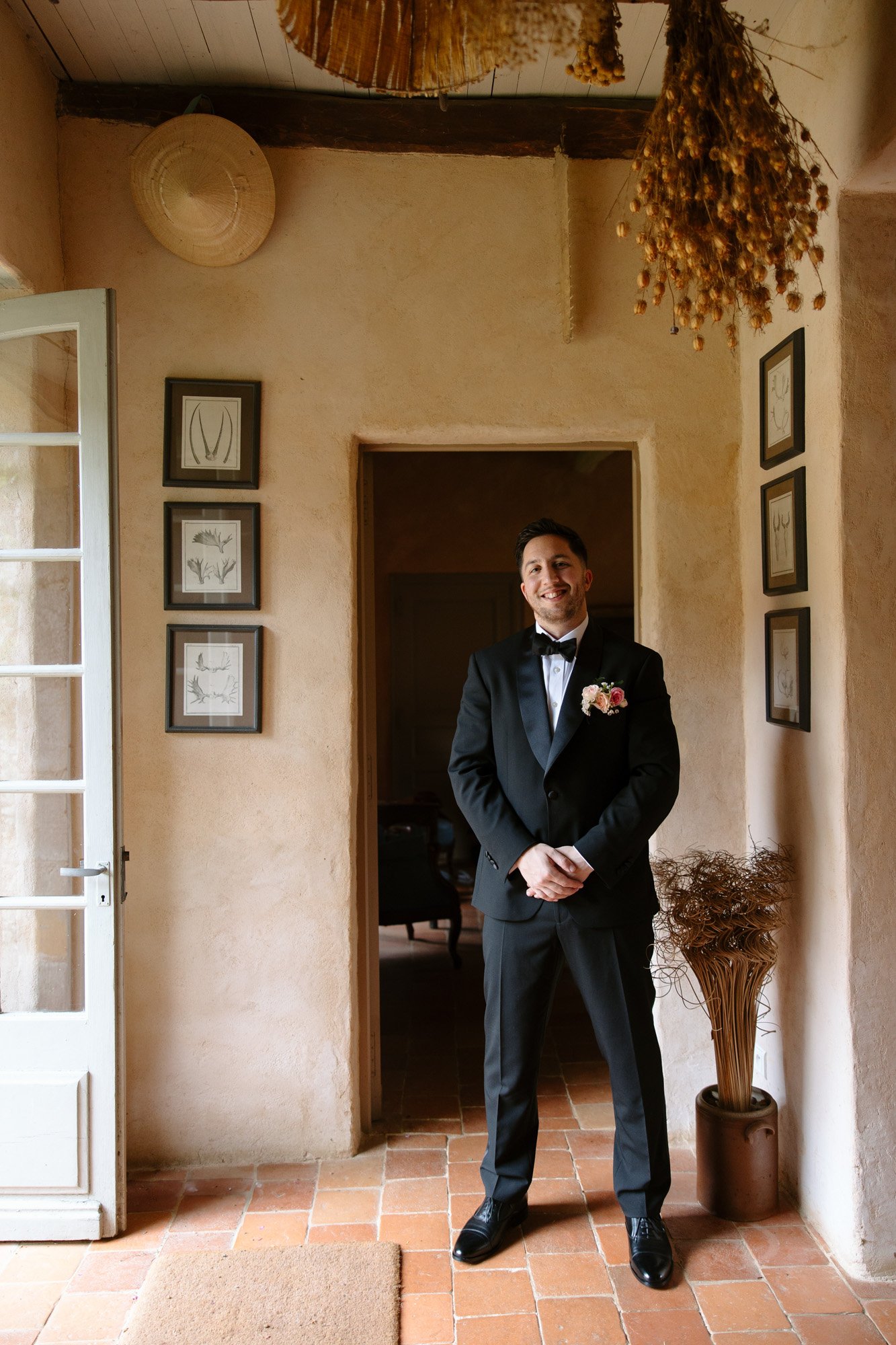 A man in a black tuxedo with a bow tie and boutonnière stands in a rustic room with framed art, tiled floor, and dried plants, smiling at the camera. Chateau de la Bourlie wedding.