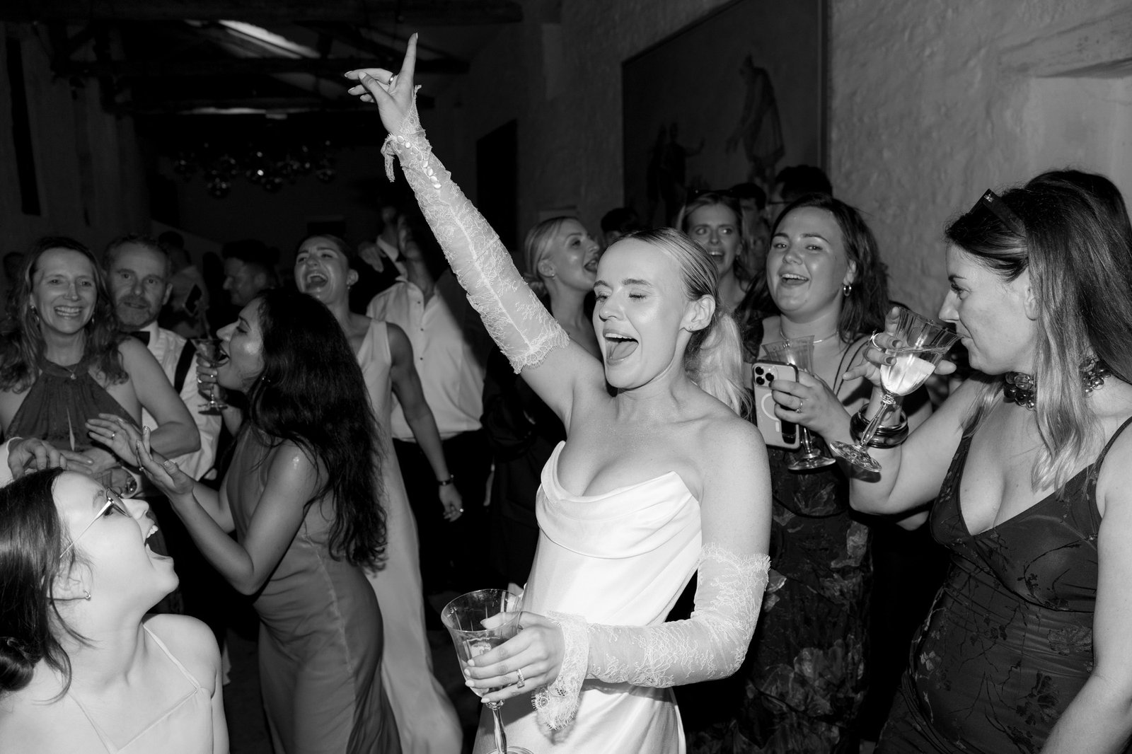 A group of people celebrate at a party, with a woman in a white dress holding a drink and raising her arm while others around her smile, laugh, and dance.