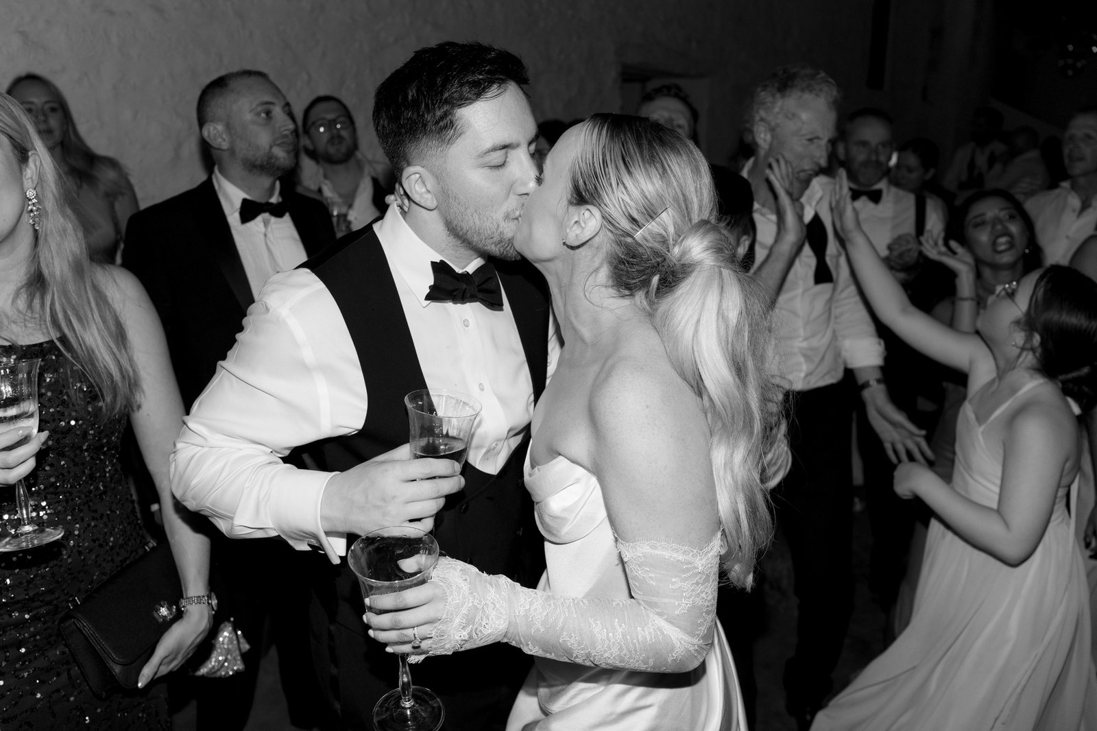 A bride and groom kiss while holding drinks at their wedding reception, surrounded by guests dressed in formal attire. Chateau de la Bourlie wedding.