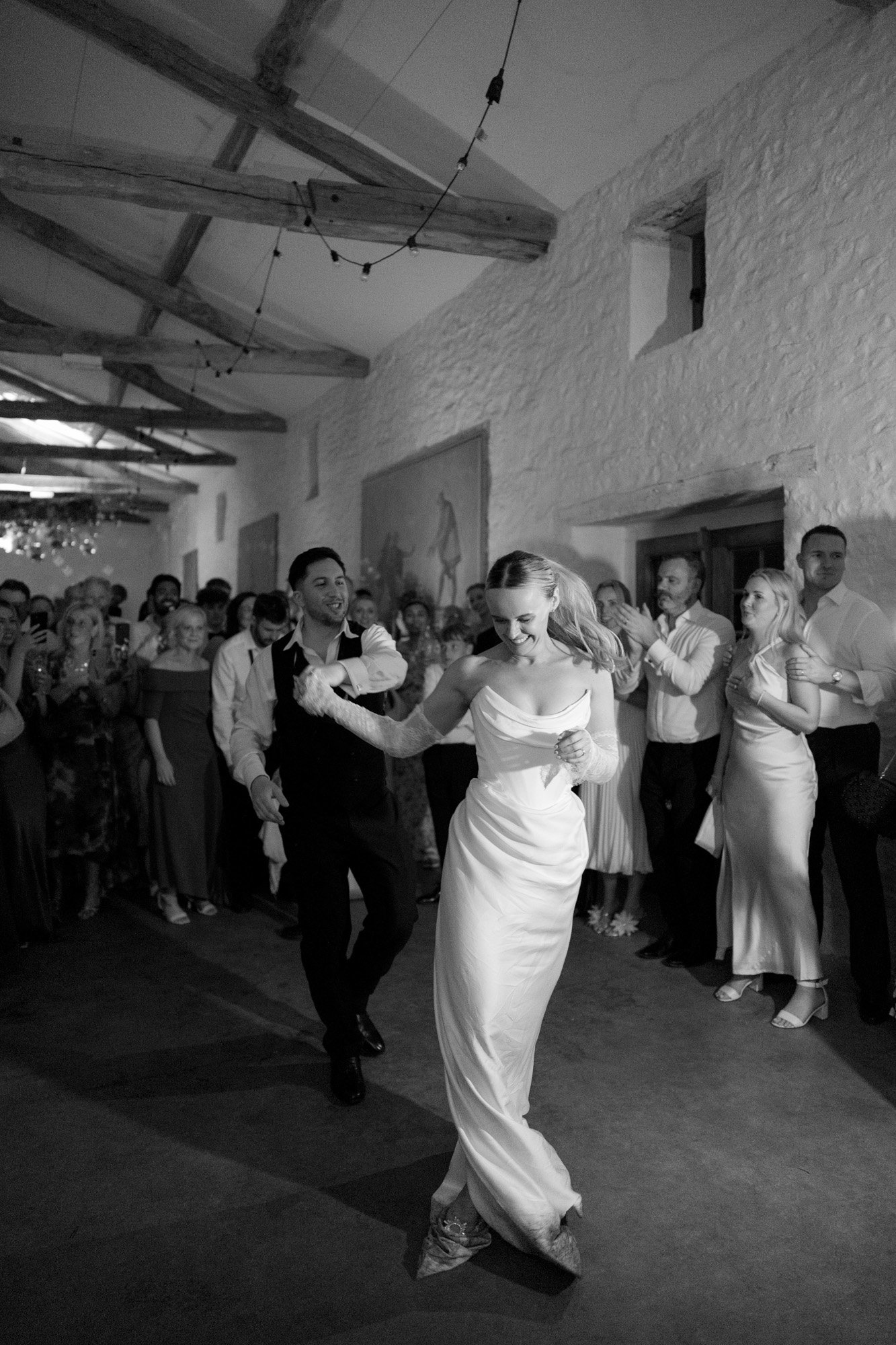 A bride and groom dance together in front of a crowd of guests inside a rustic venue with exposed beams and stone walls. Chateau de la Bourlie wedding.