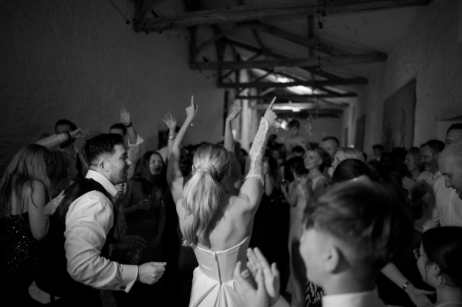 A crowd of people, including a bride and groom, dance and celebrate together indoors under exposed wooden beams. Chateau de la Bourlie wedding.