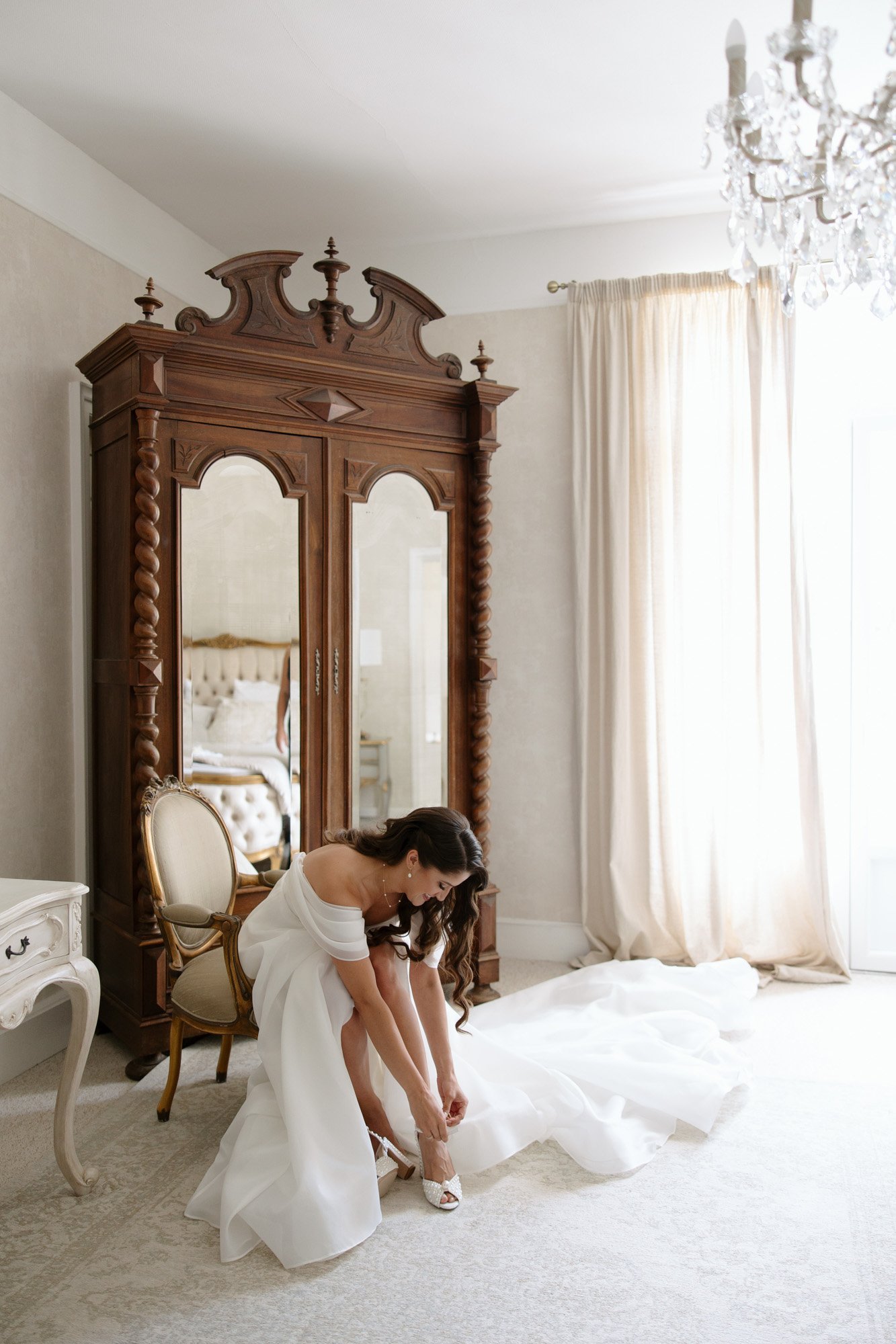 A bride in a white dress bends down to fasten her shoe in a bright, elegant room with a large wooden wardrobe and a chandelier. Chateau de la Couronne wedding.