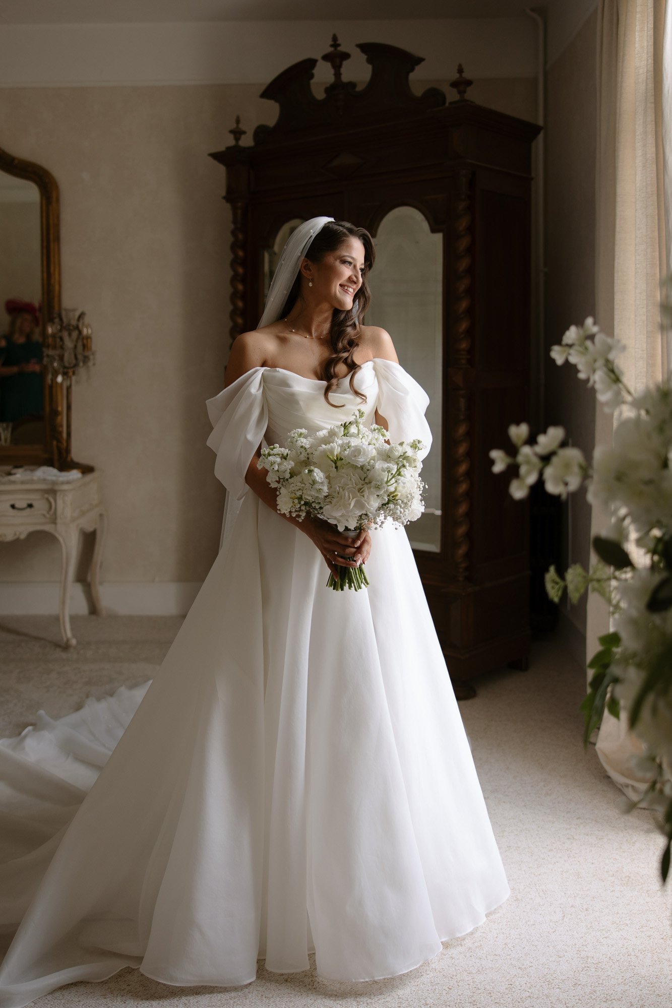 A bride in an off-the-shoulder white gown holds a bouquet of white flowers and stands near a window, smiling, in a softly lit room with classic decor. Chateau de la Couronne wedding.