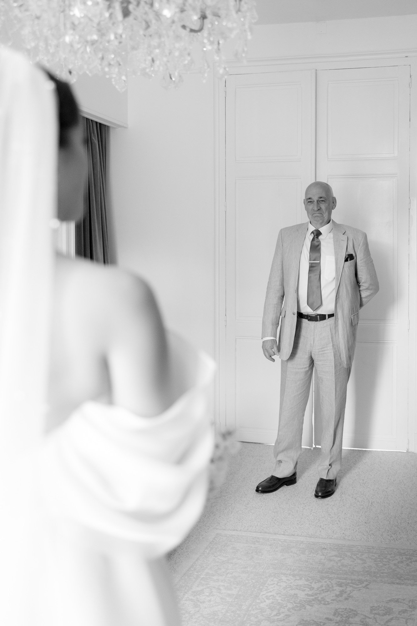 A man in a light-colored suit stands facing a woman in a wedding dress indoors, with a chandelier visible above.