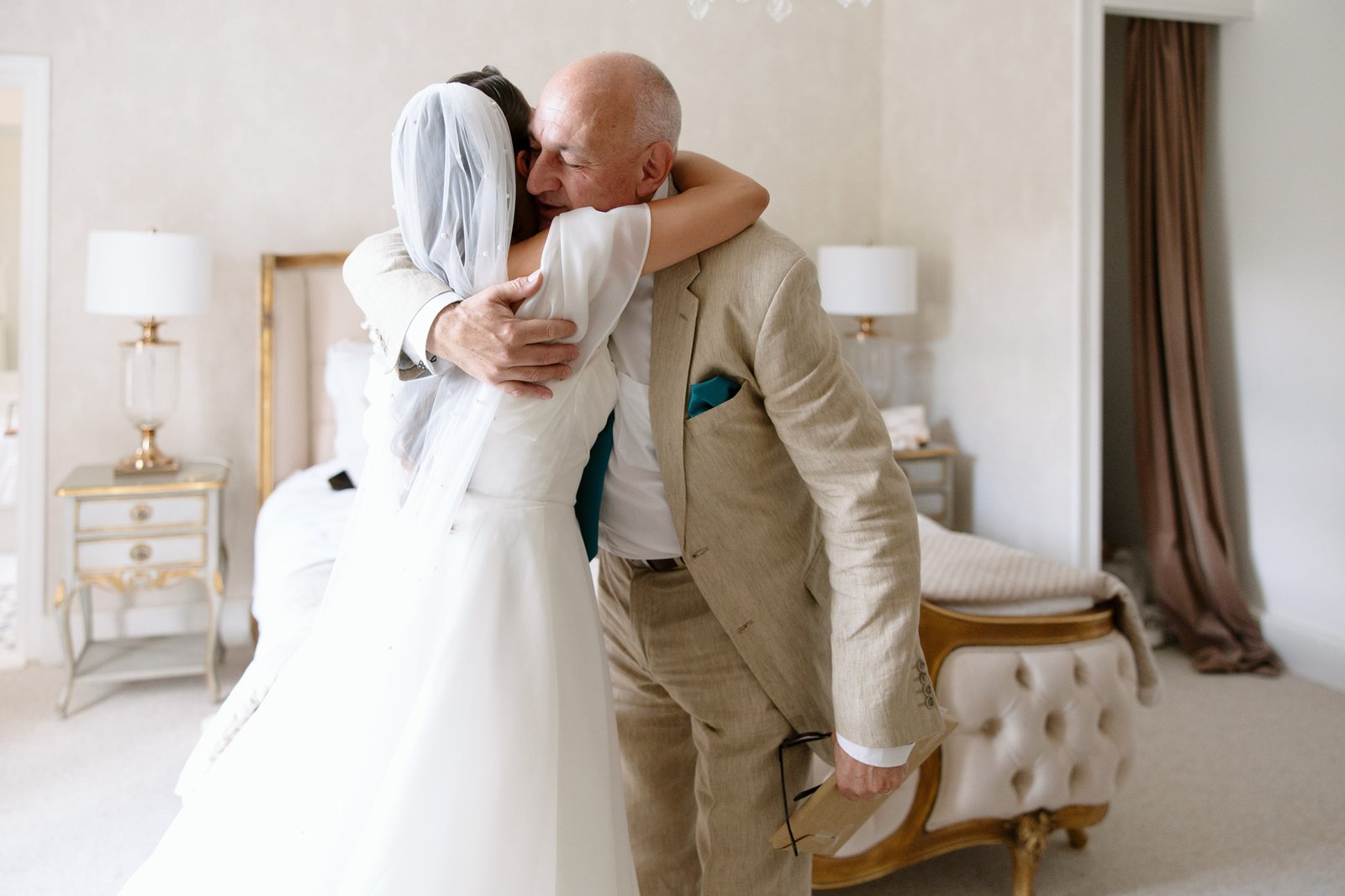 A bride in a white dress and veil hugs an older man in a beige suit in a bedroom with elegant furniture. Chateau de la Couronne wedding.