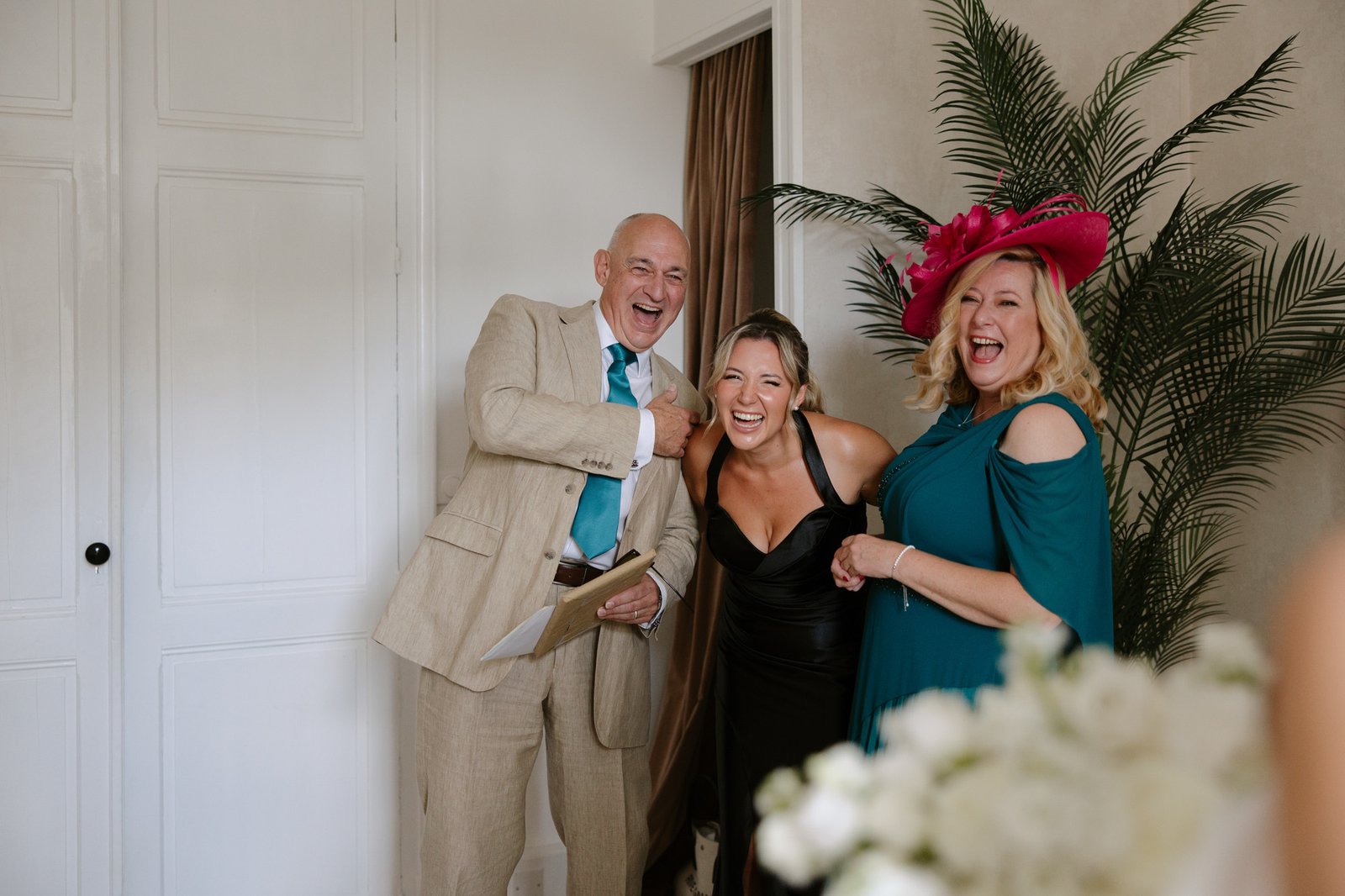 Three adults dressed formally stand indoors, smiling and laughing. One man wears a tan suit, and two women wear elegant dresses; one has a large pink hat. A large plant decorates the background.