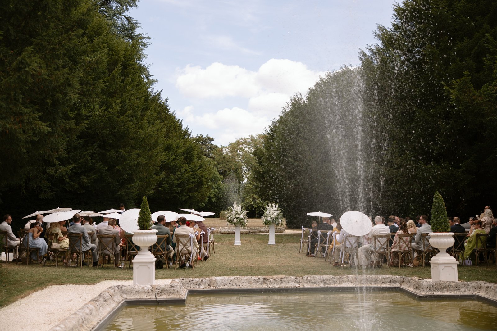 Outdoor wedding ceremony with guests seated on either side of an aisle, some holding white parasols, near a fountain and surrounded by tall trees. Chateau de la Couronne wedding.