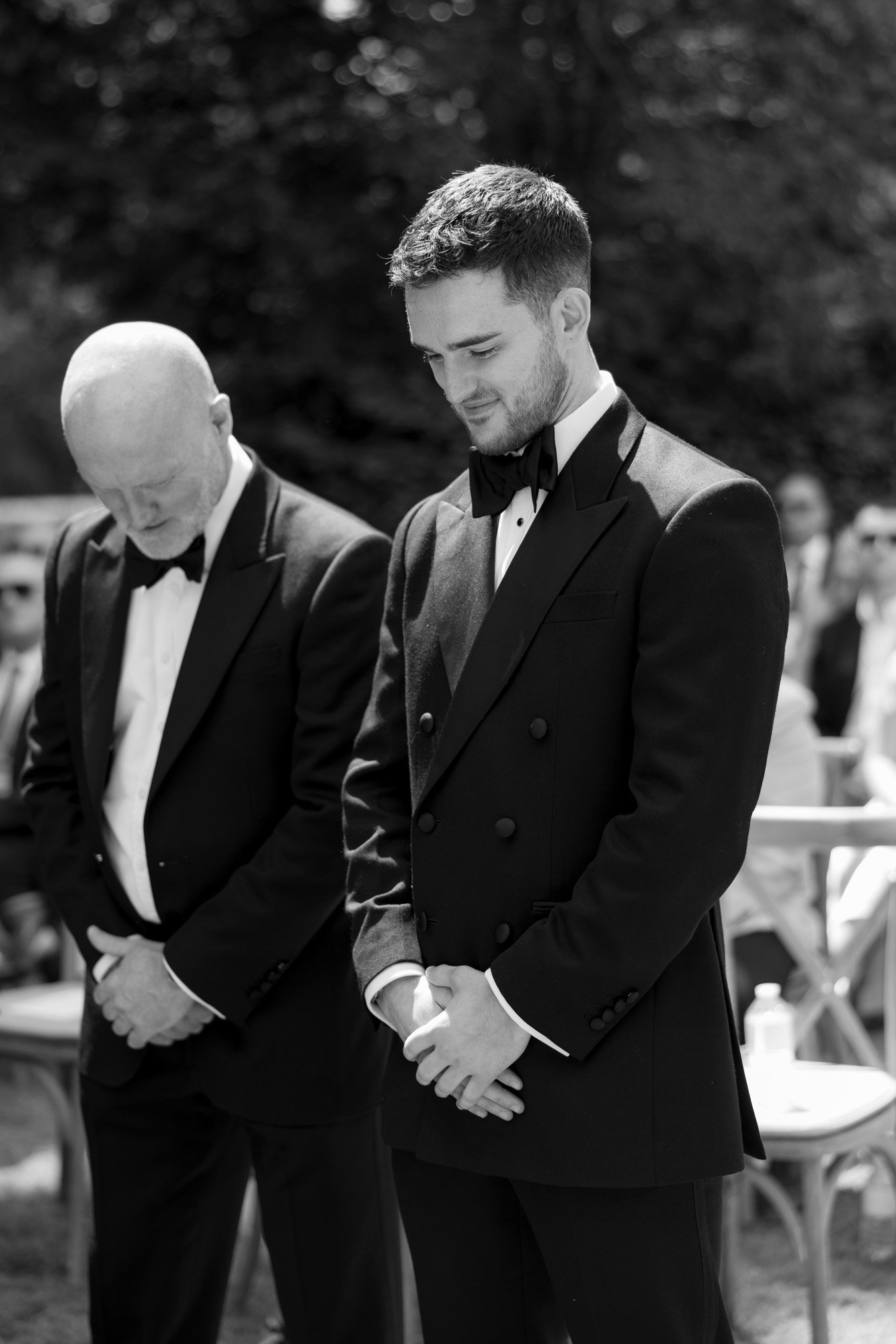 Two men in tuxedos stand outdoors with heads bowed, appearing solemn or reflective, during a formal event with people seated in the background.
