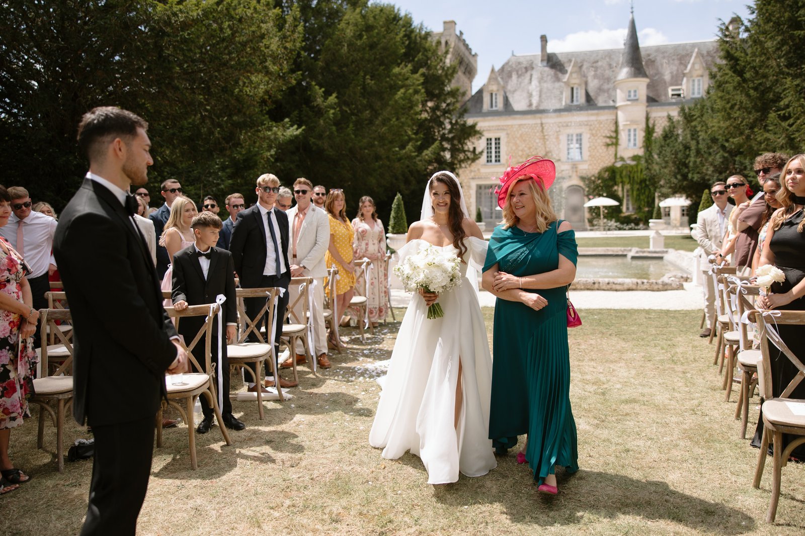 A bride holding a bouquet walks down an outdoor aisle with a woman in a teal dress and pink hat, as guests look on at a wedding ceremony. Chateau de la Couronne wedding.
