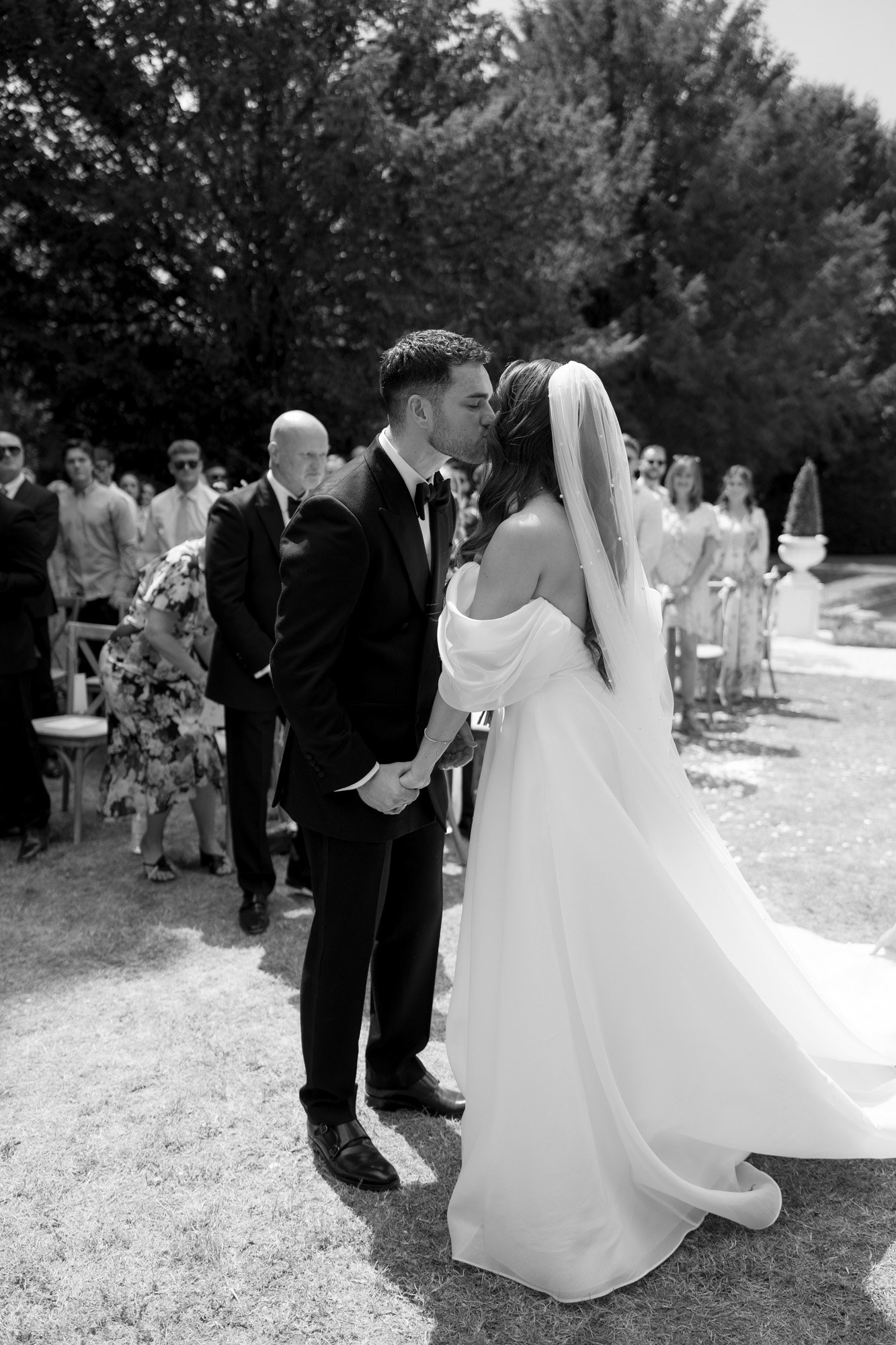 A bride and groom kiss outdoors during their wedding ceremony, holding hands while guests watch in the background. Chateau de la Couronne wedding.