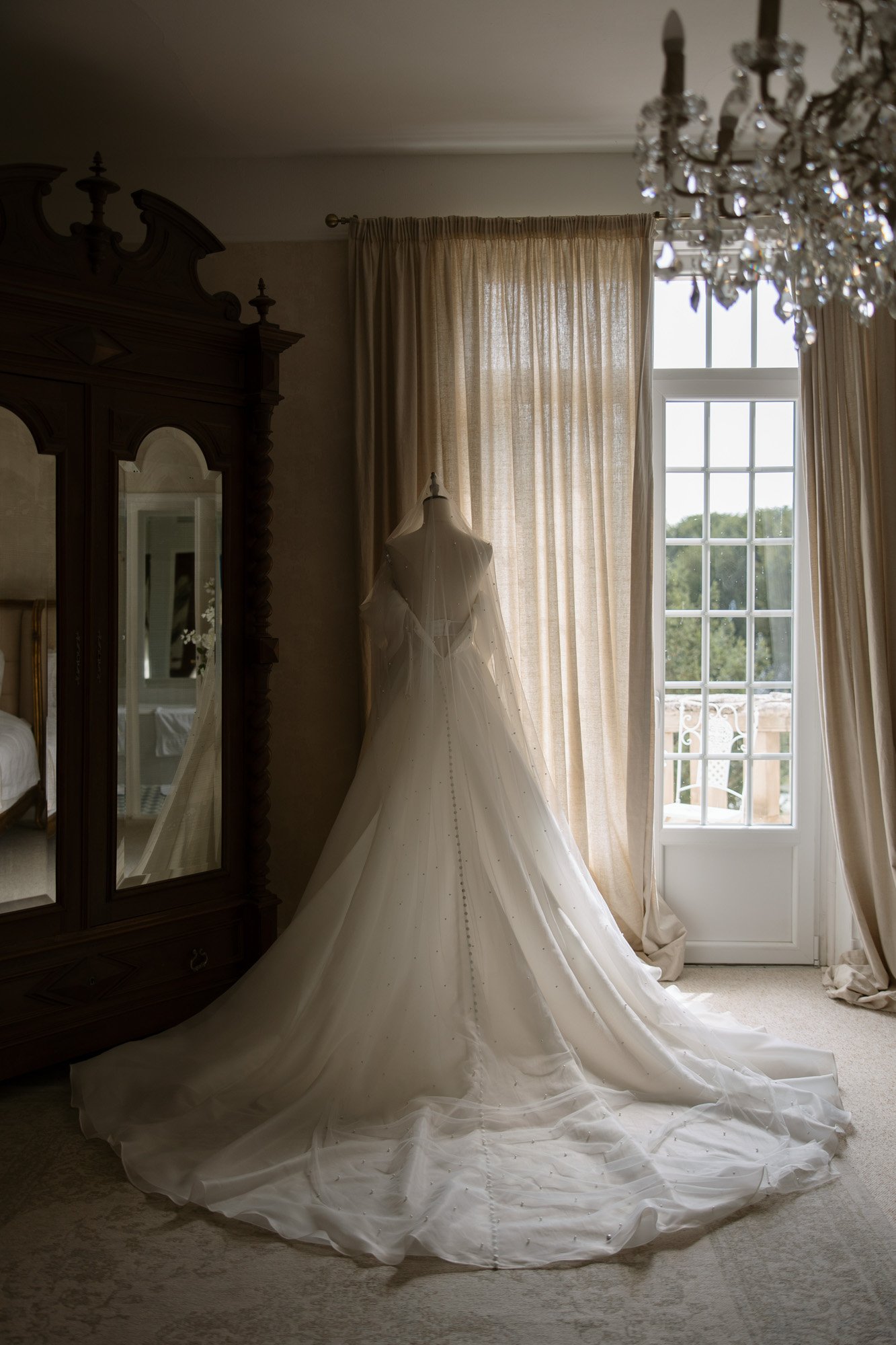 A white wedding dress displayed on a mannequin stands in front of a window with sheer curtains, beside a wooden wardrobe and a chandelier above.