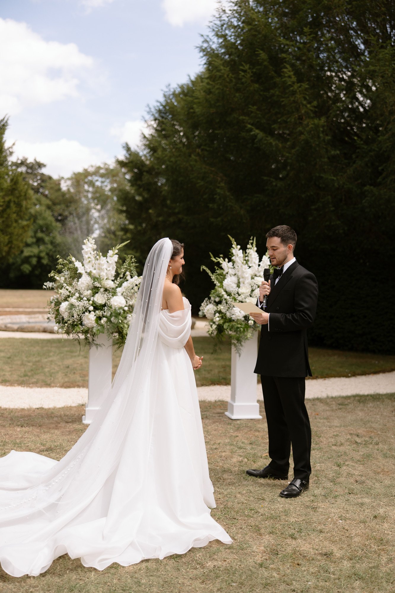 A bride and groom stand outdoors facing each other; the groom holds a microphone, reciting vows. Two large floral arrangements are behind them.