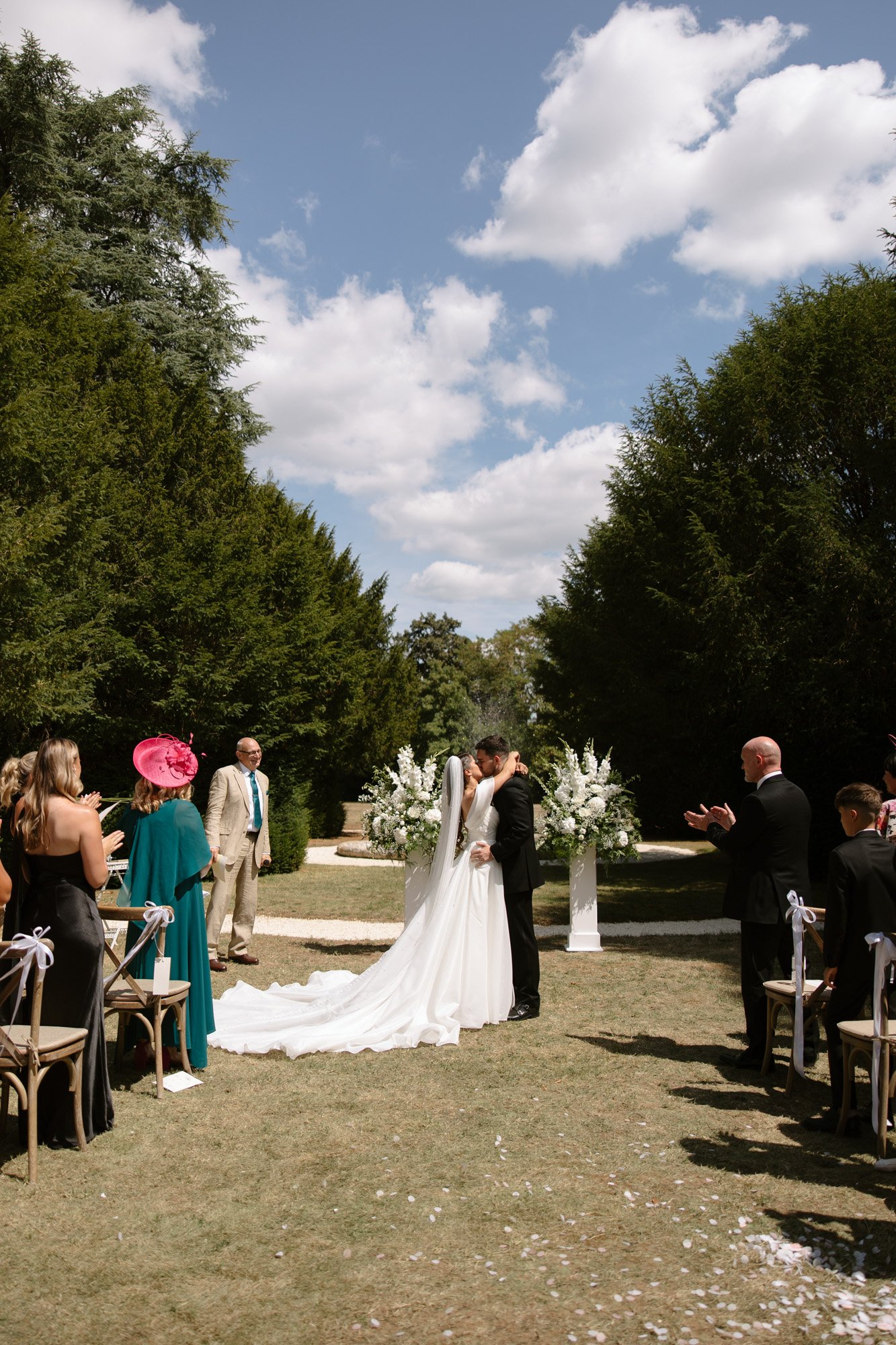 A bride and groom kiss at an outdoor wedding ceremony under a blue sky, surrounded by guests and floral arrangements. Chateau de la Couronne wedding.