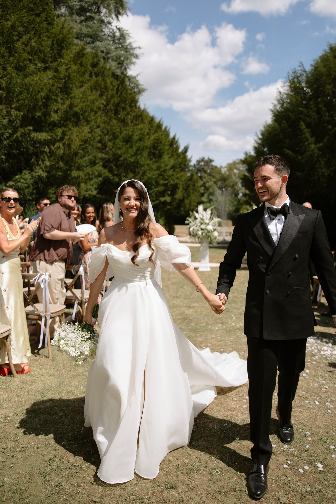 A bride and groom walk hand in hand outdoors, smiling, as guests seated on wooden chairs clap and celebrate under a sunny sky. Chateau de la Couronne wedding.