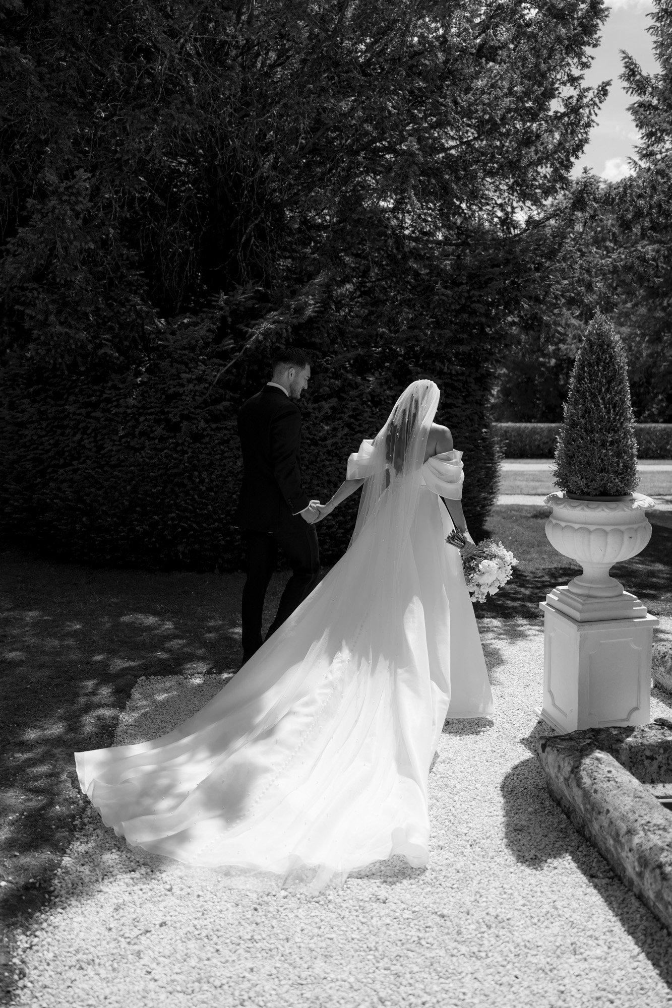 A bride and groom walk hand in hand outdoors; the bride wears a long white gown and veil, and holds a bouquet. The scene is in black and white. Chateau de la Couronne wedding.