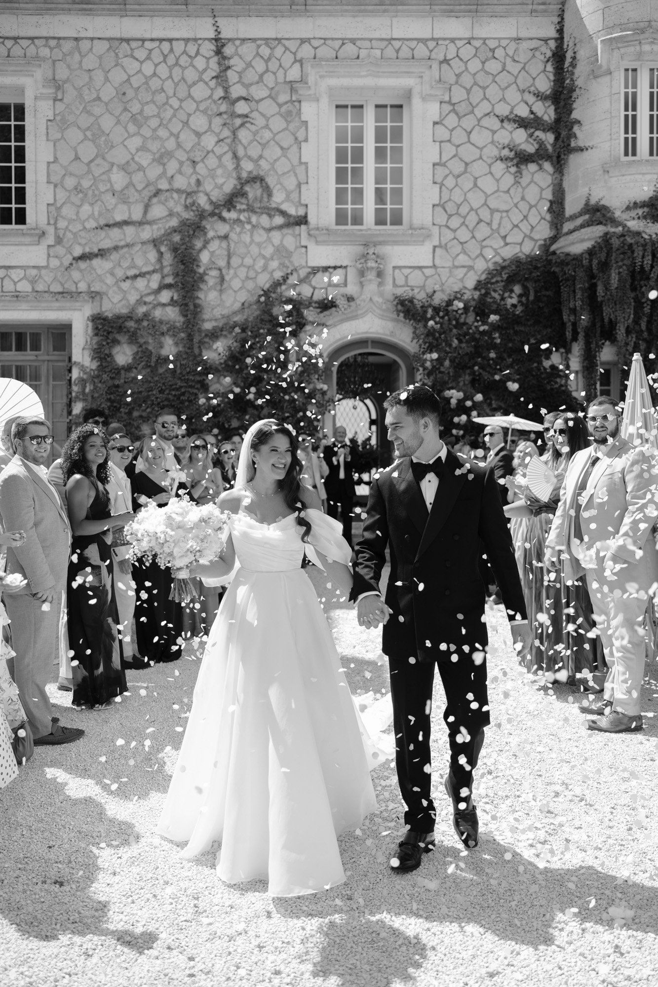 A bride and groom walk hand in hand outside a building as guests throw flower petals in celebration, with people smiling and dressed formally. Chateau de la Couronne wedding.