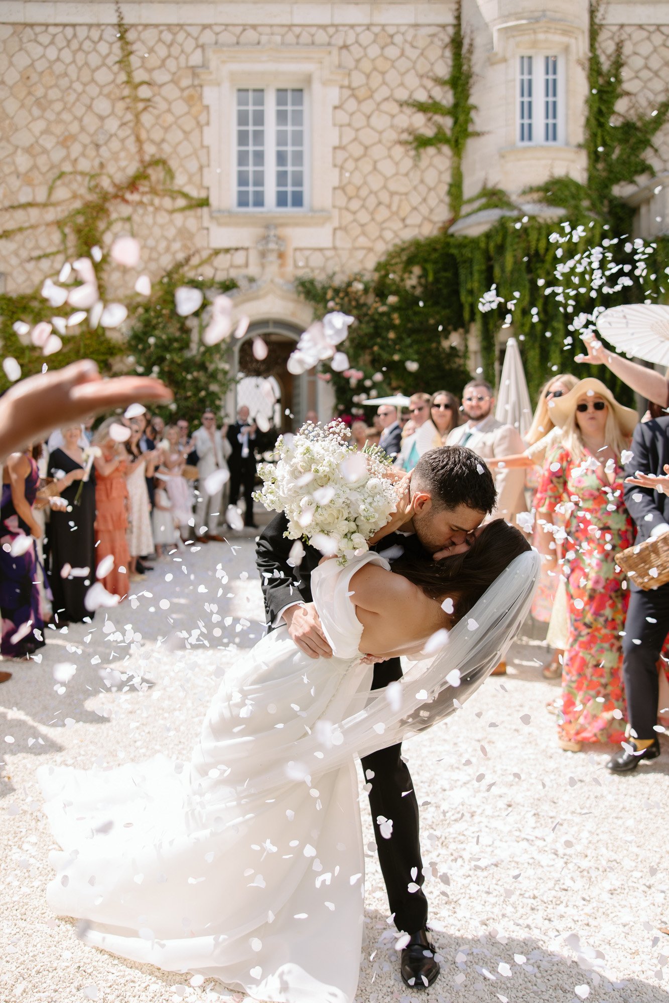 A bride and groom kiss as the groom dips the bride, surrounded by guests throwing flower petals outside a stone building during a wedding celebration.