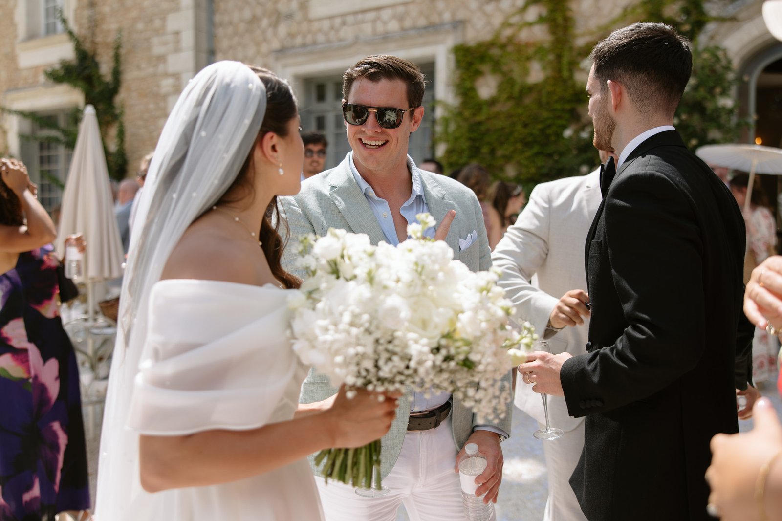 A bride holding a white bouquet talks with guests, including a man in sunglasses, at an outdoor wedding reception.